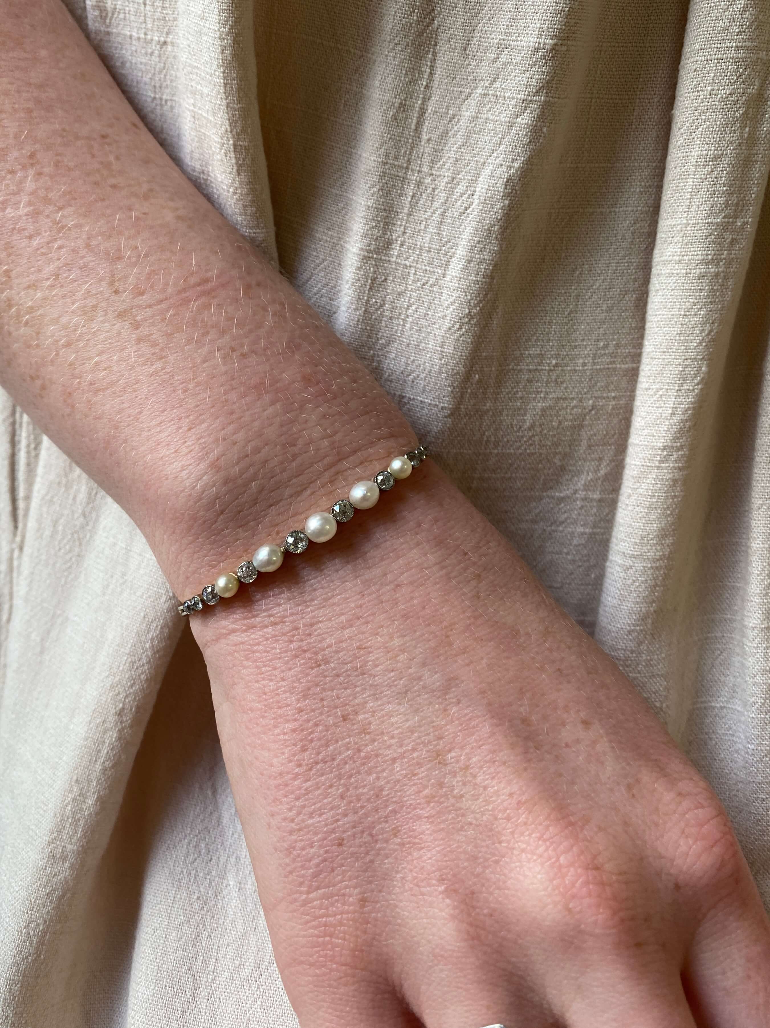 A close-up of a gold bracelet with white pearls and old cut diamonds worn on a person's wrist and shown against a soft beige linen background.