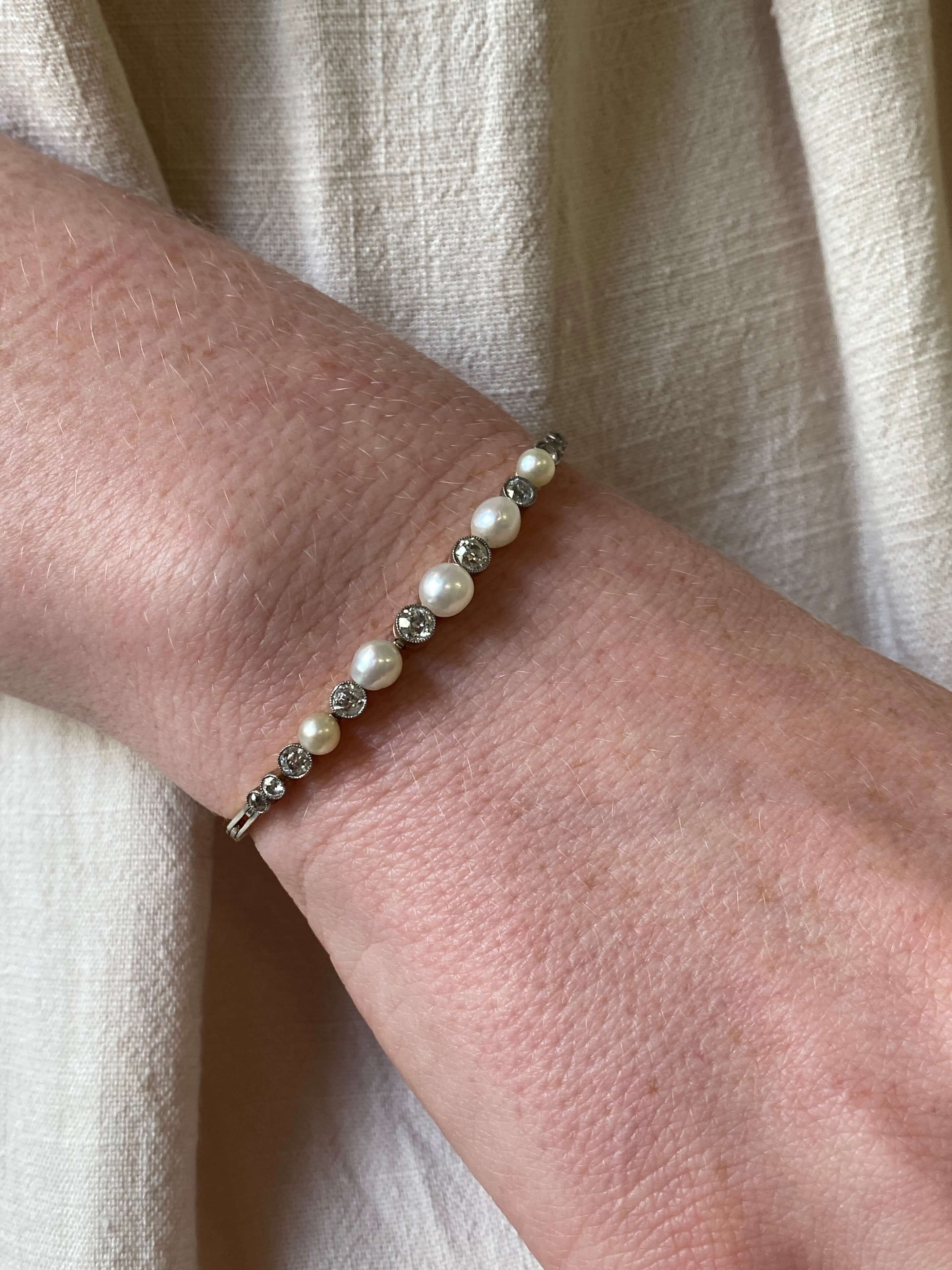 A close-up of a gold bangle bracelet with white pearls and old cut diamonds worn on a person's wrist and set against a soft beige linen fabric background.