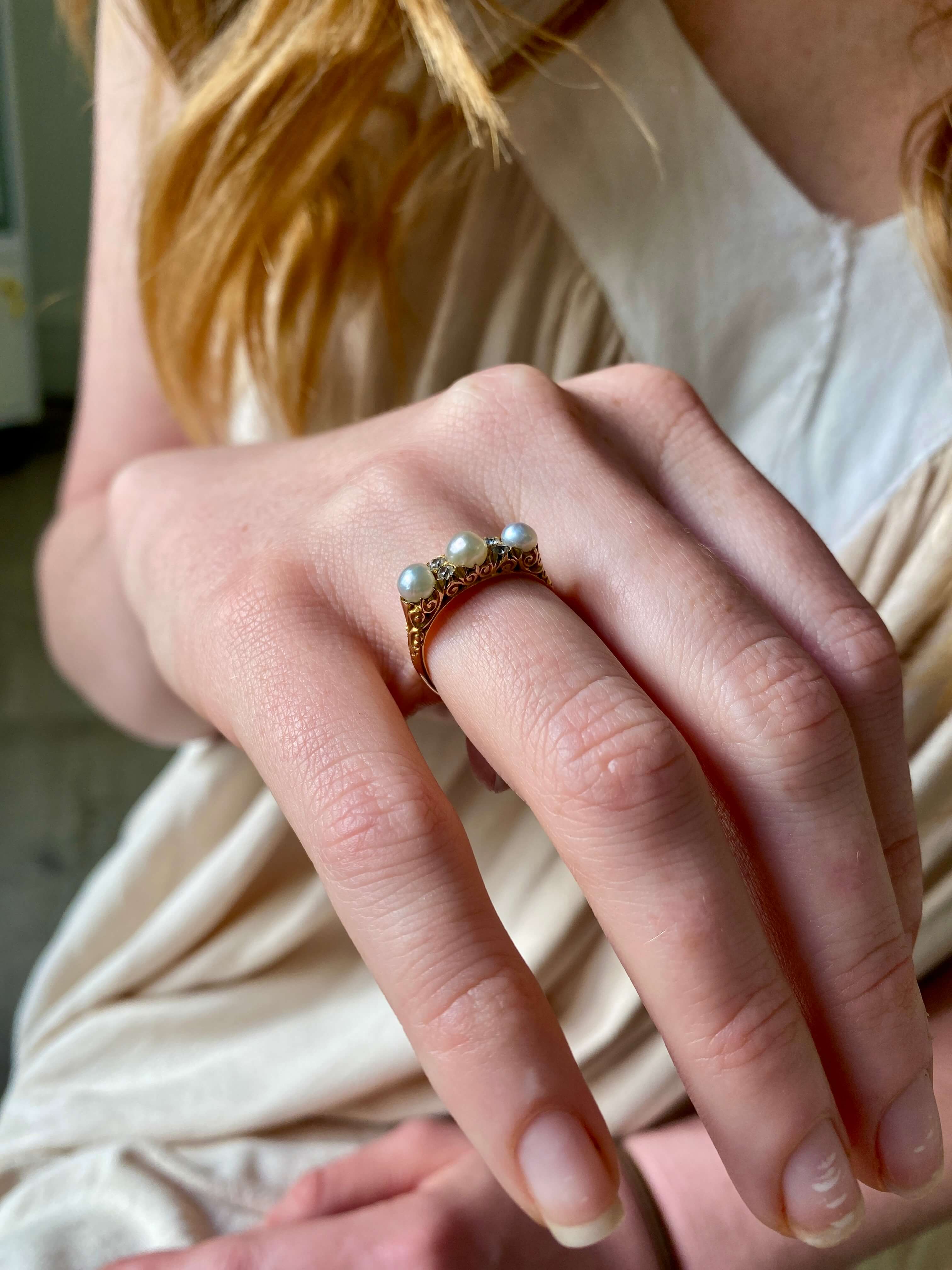 A close-up of a person's hand wearing a yellow gold ring with three small white pearls, set against a soft cream-colored dress.