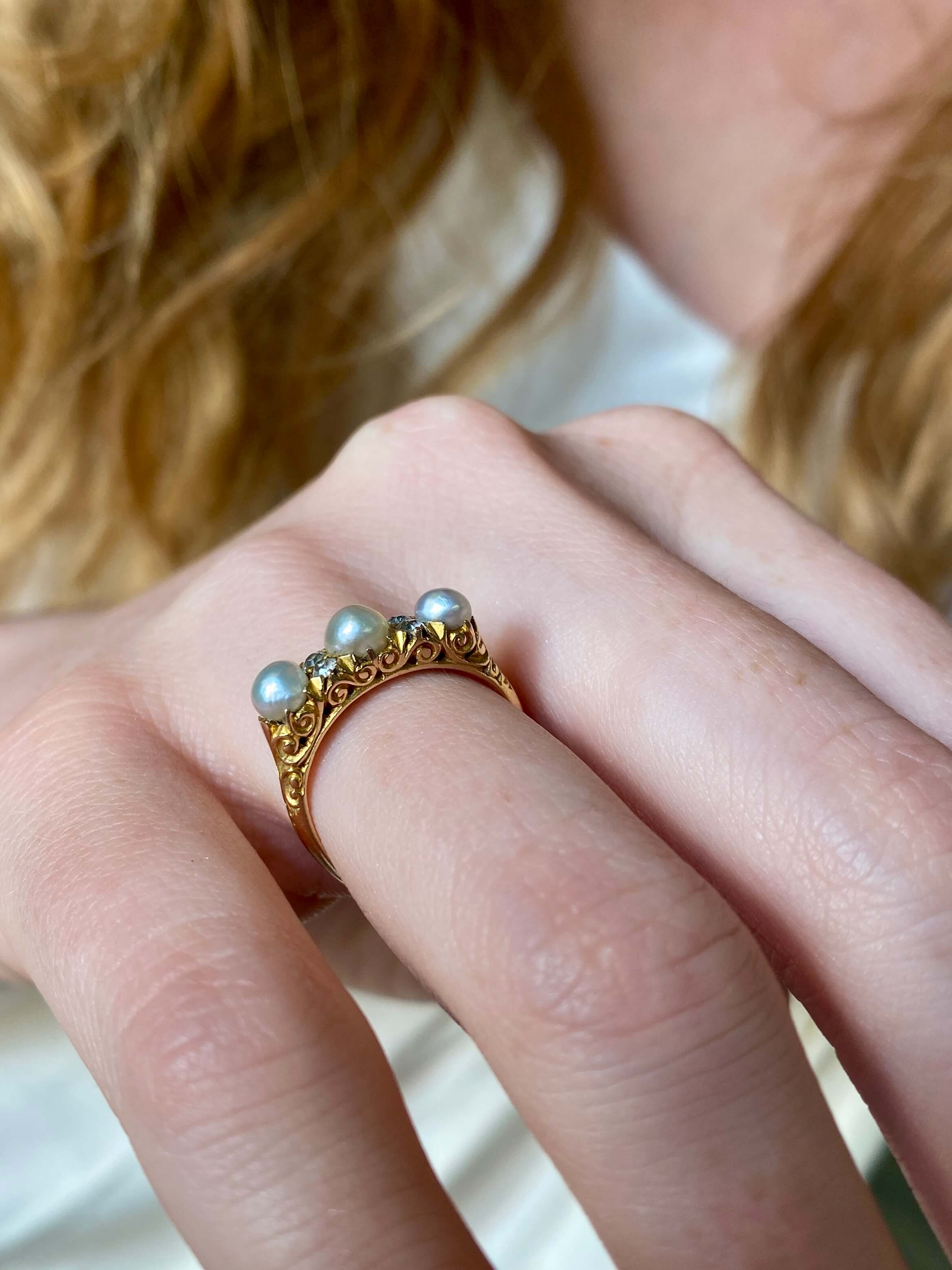 A close-up of a person's hand wearing a vintage-style gold ring adorned with three small round pearls and delicate filigree detailing.