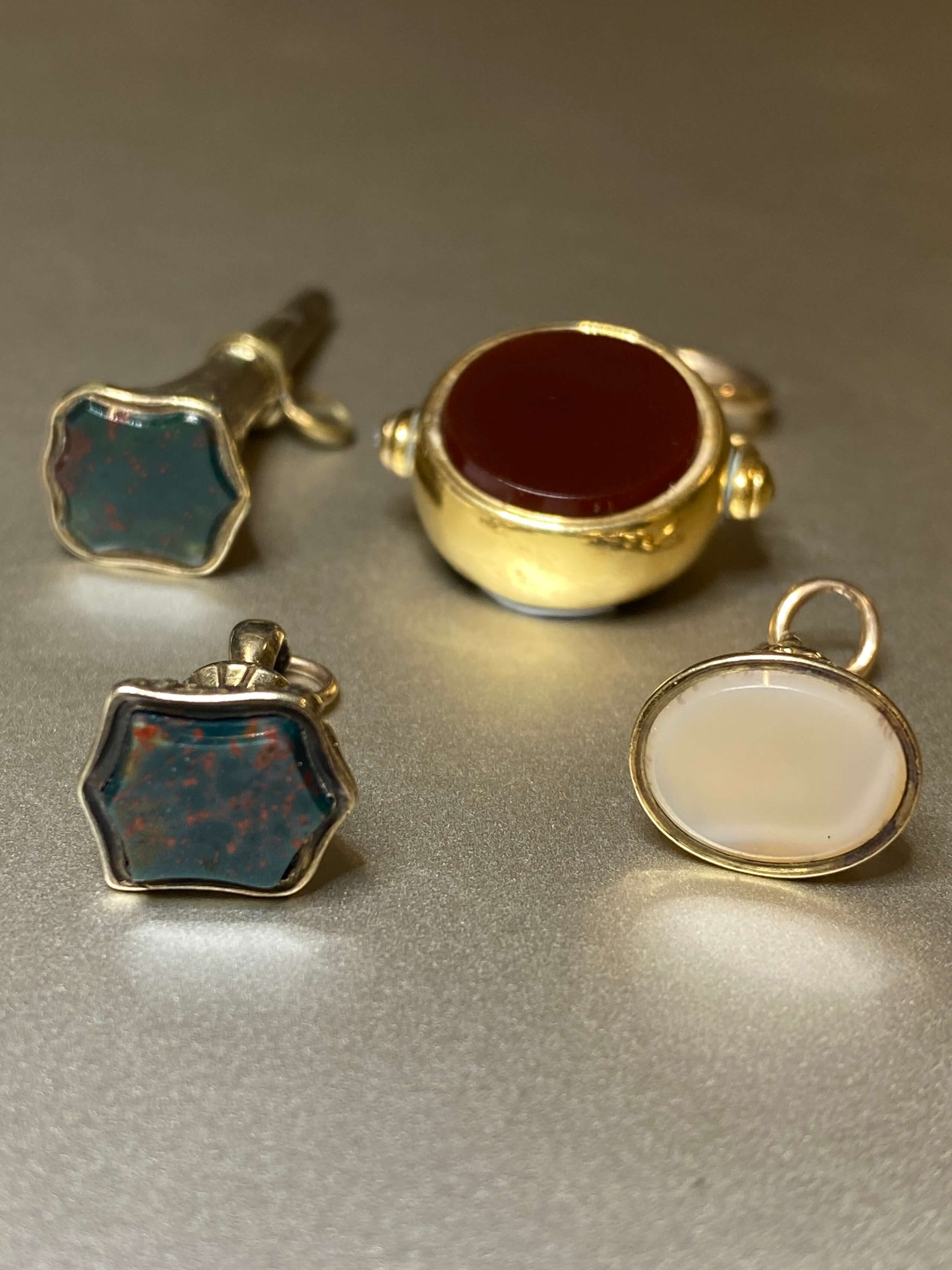 Four vintage gold-toned jewelry pieces displayed on a neutral gray surface: a hexagonal stud earring with a blue-green stone, a hexagonal stud earring with a red-orange stone, a gold signet ring with a deep red stone, and an oval bezel setting with a milky white stone.