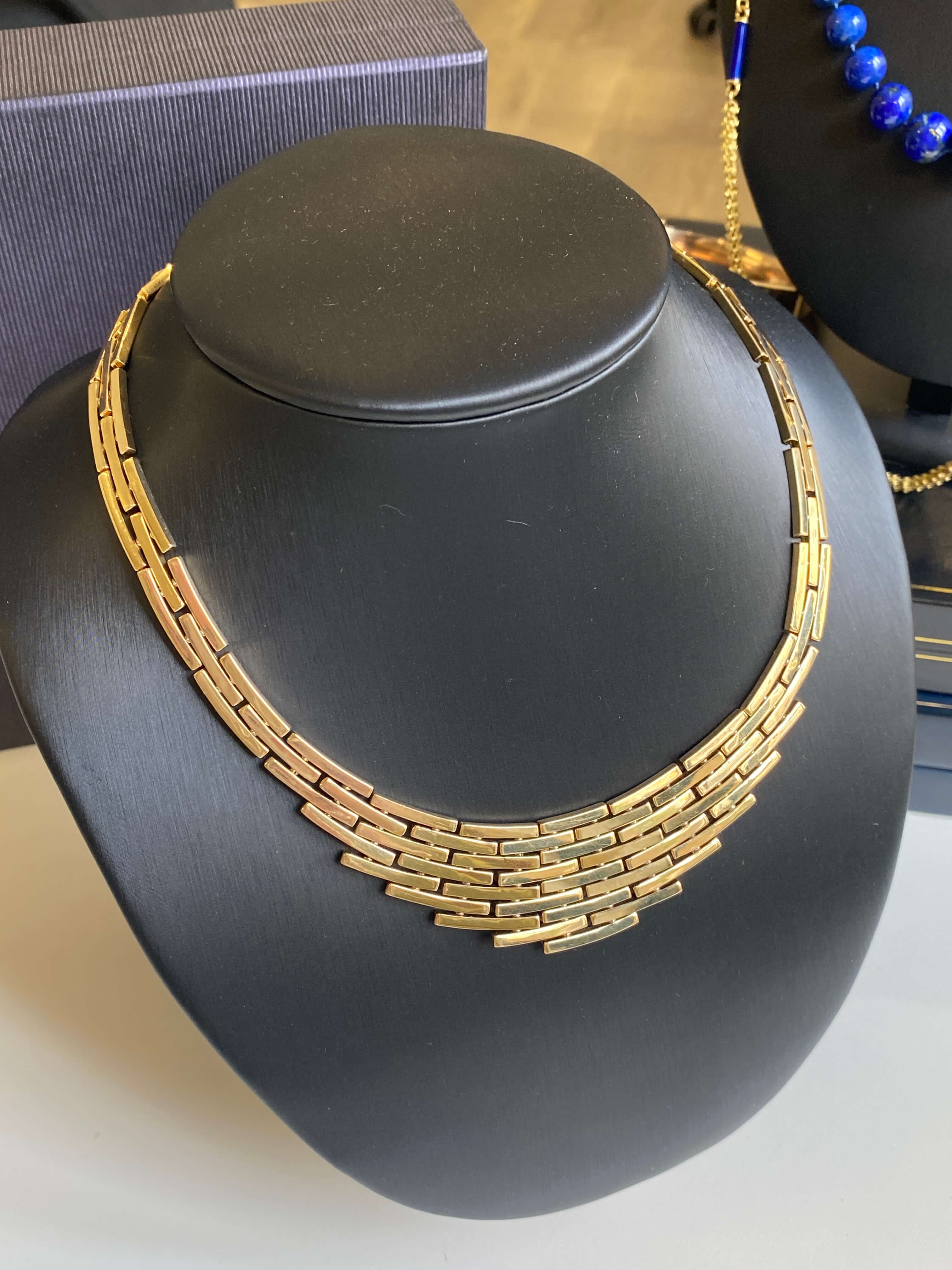 A gold-toned multi-strand chain necklace displayed on a black jewelry bust, with part of a blue beaded necklace visible in the background.