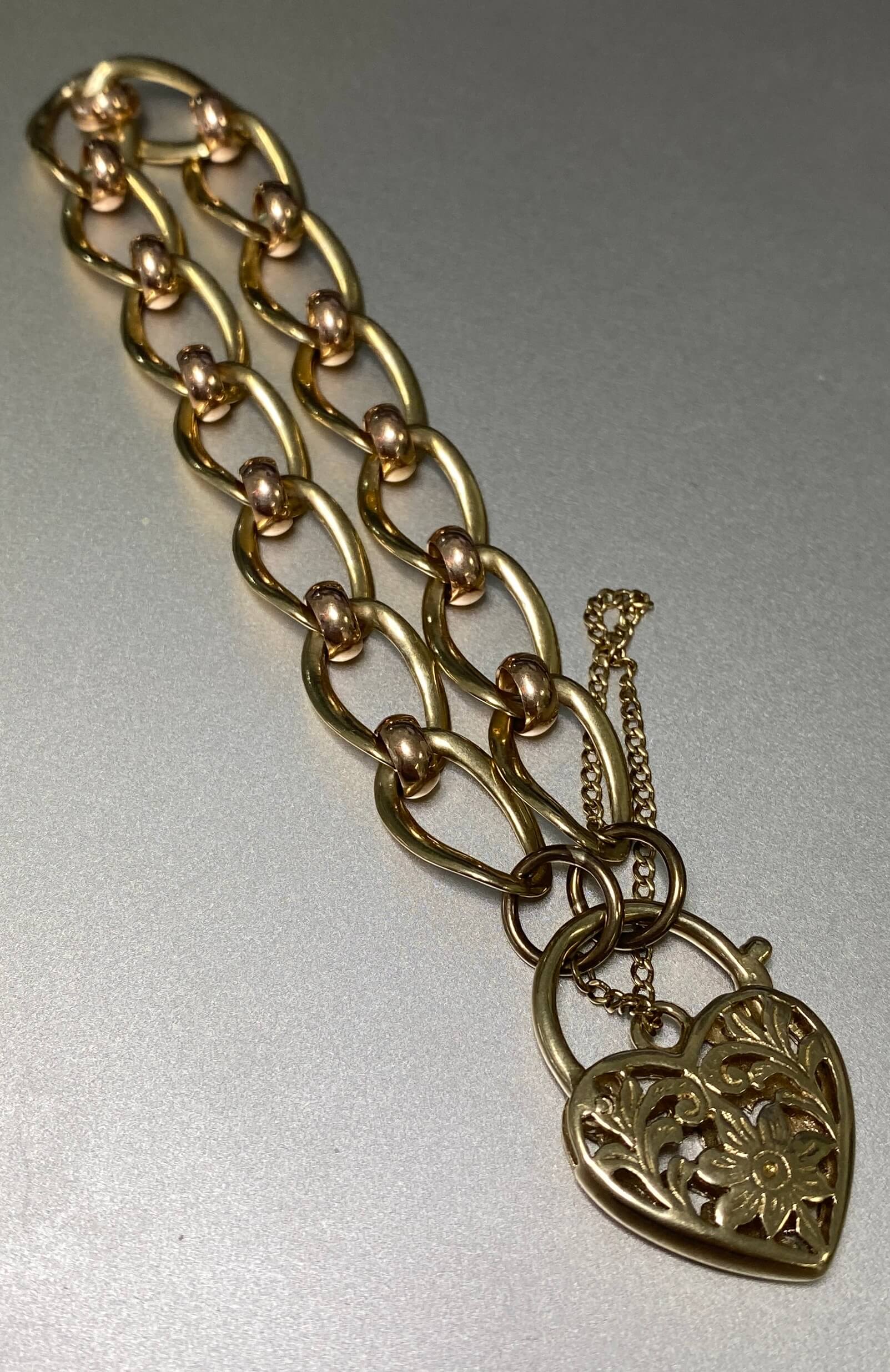 A yellow and rose gold bracelet featuring a large interlocking link chain and a heart-shaped padlock pendant with floral cutout detailing, photographed on a plain light gray background.