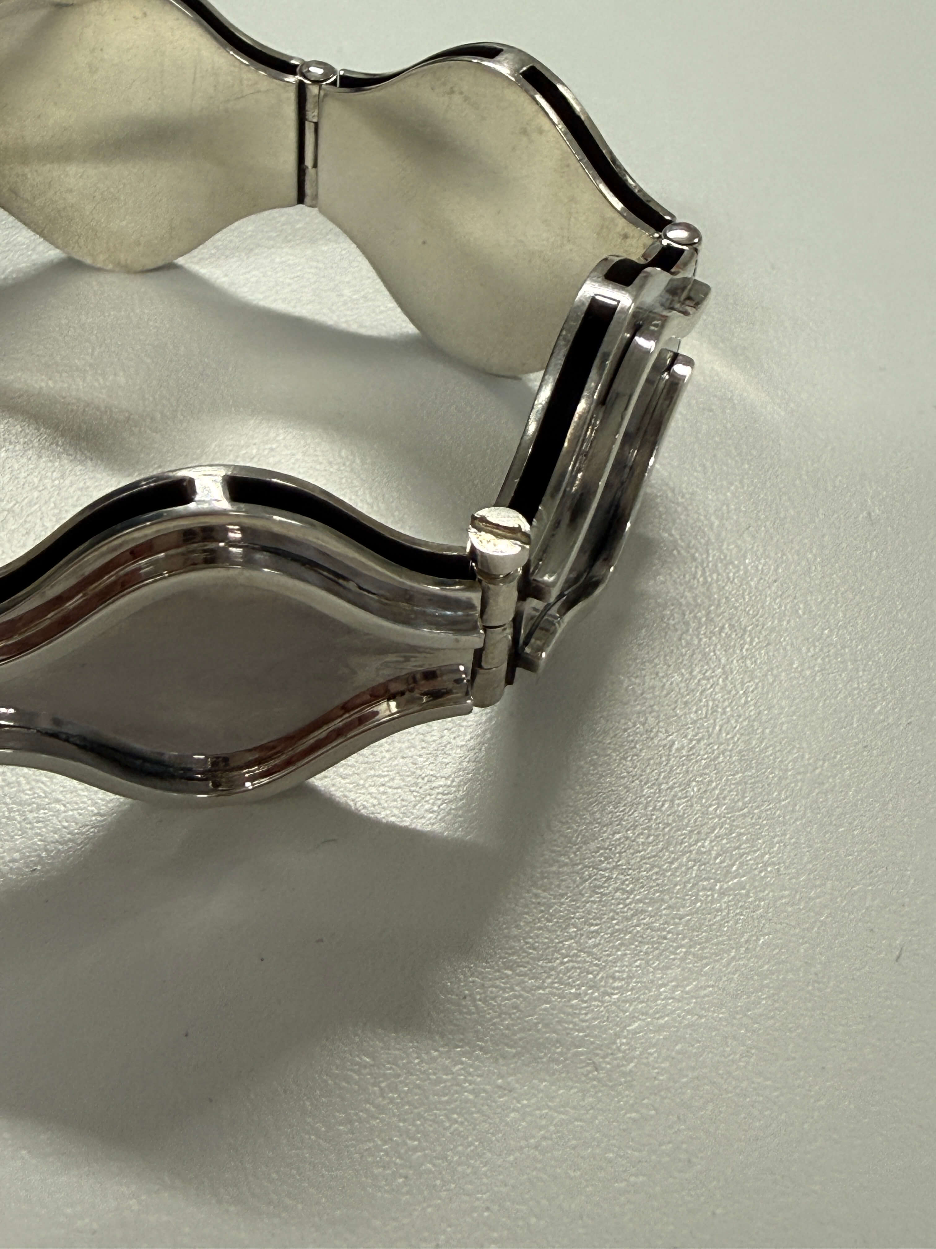 A close-up of a polished silver-tone metal bracelet with a wavy, scalloped edge design, resting on a light gray textured surface.