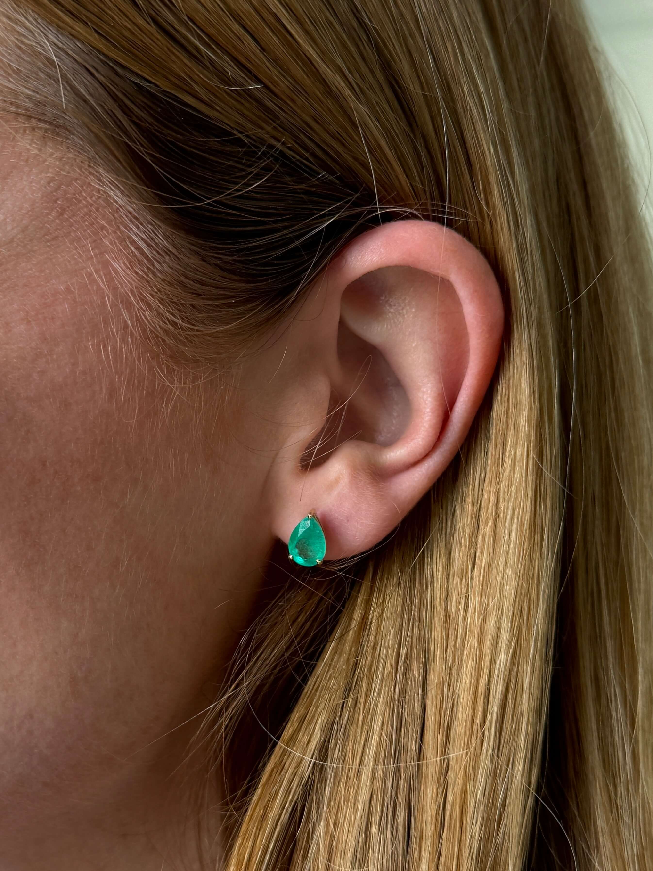 A close-up of a woman's ear wearing a small pear-shaped emerald stud earring.