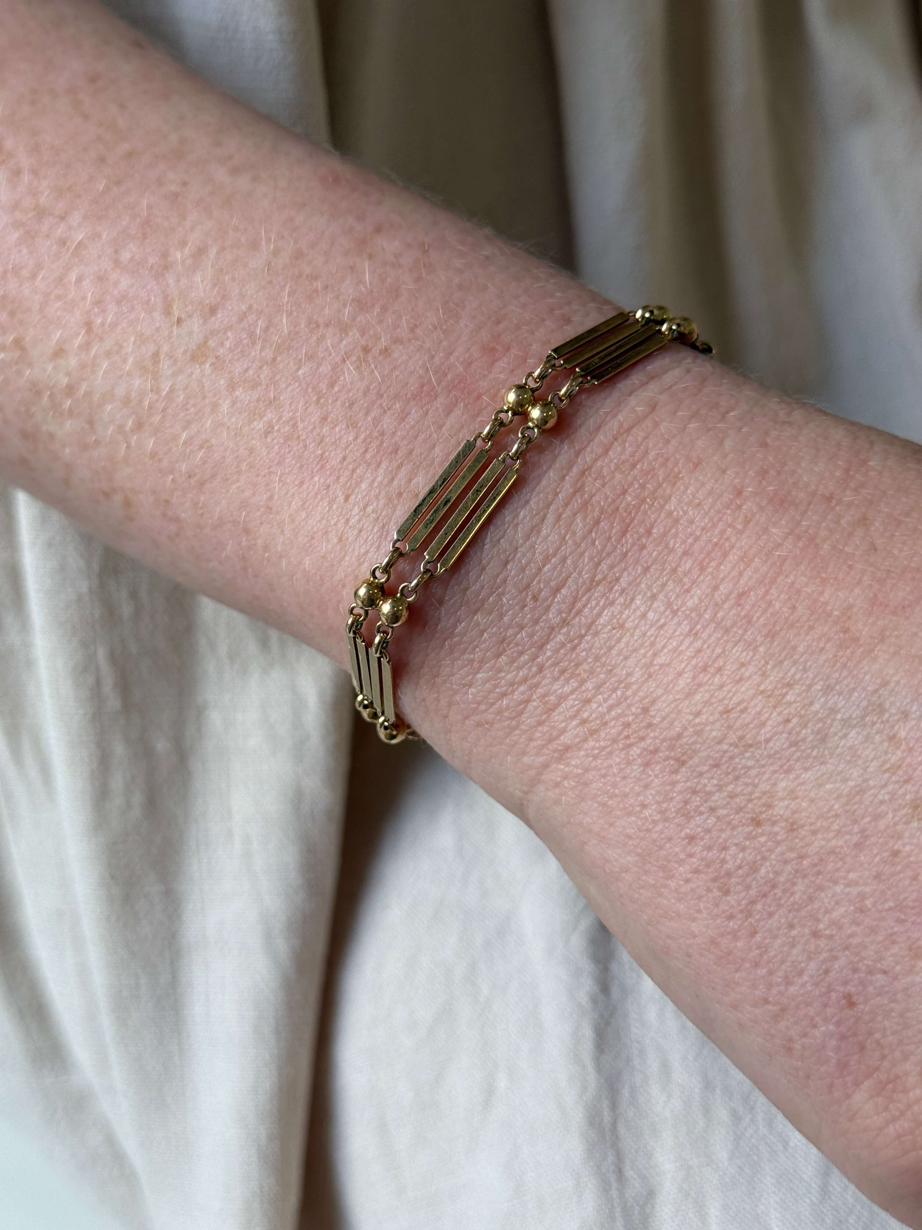 A close-up of a person's wrist wearing a delicate gold-toned bracelet with elongated rectangular links and small bead details, set against a soft beige fabric background.