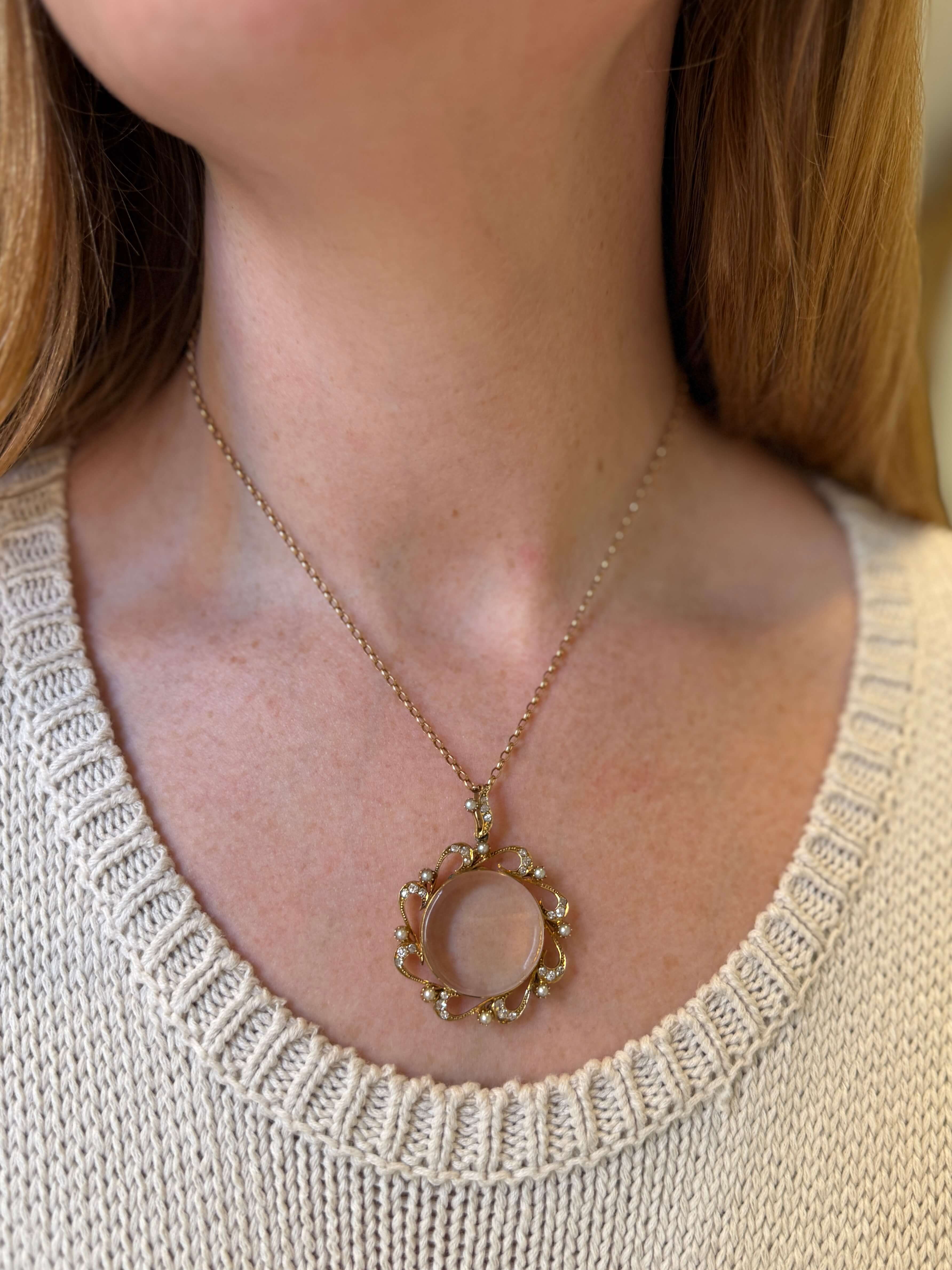 A close-up of a woman wearing a delicate gold necklace with a circular pearl & diamond pendant featuring a pale pink stone in the center, paired with a cream-colored knit sweater.