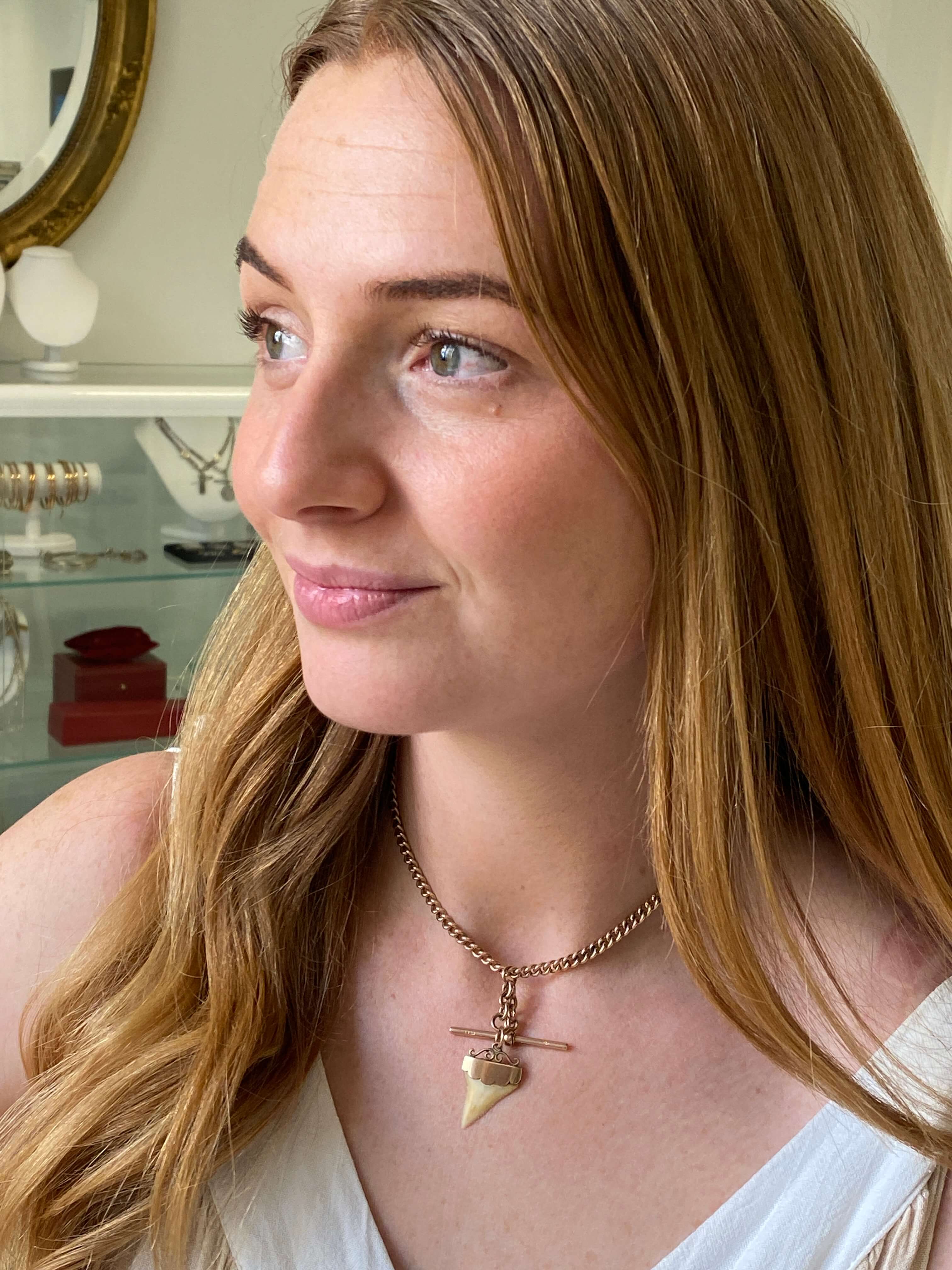 A close-up lifestyle shot of a woman with long light brown hair wearing a gold chain necklace with a shark tooth pendant in a jewelry store setting.
