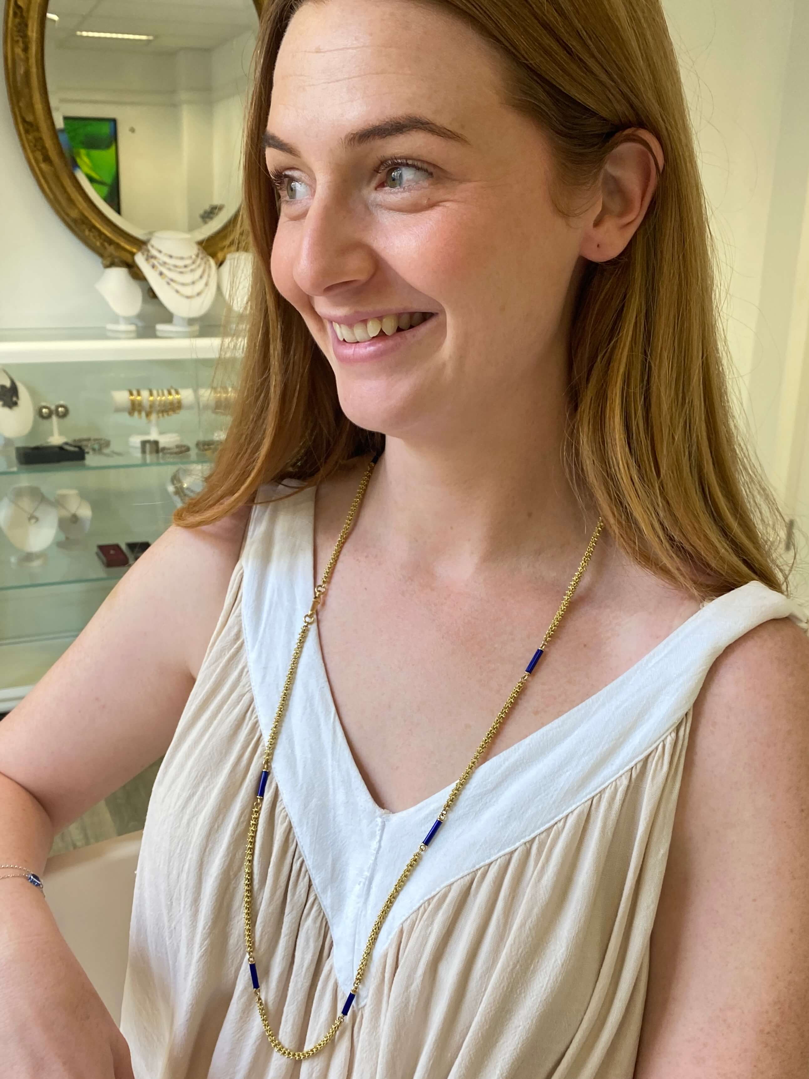 A woman with long hair, wearing a long gold chain necklace with blue enamel accents. 