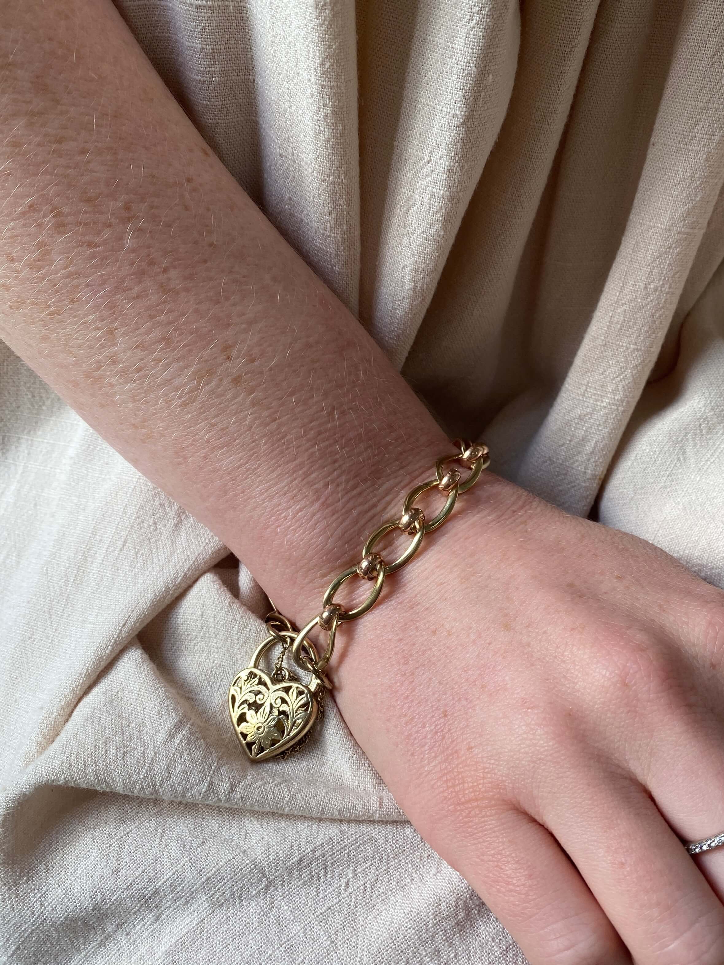 A close-up of a person's wrist wearing a gold chain bracelet with a heart-shaped padlock charm, set against a background of draped beige fabric.