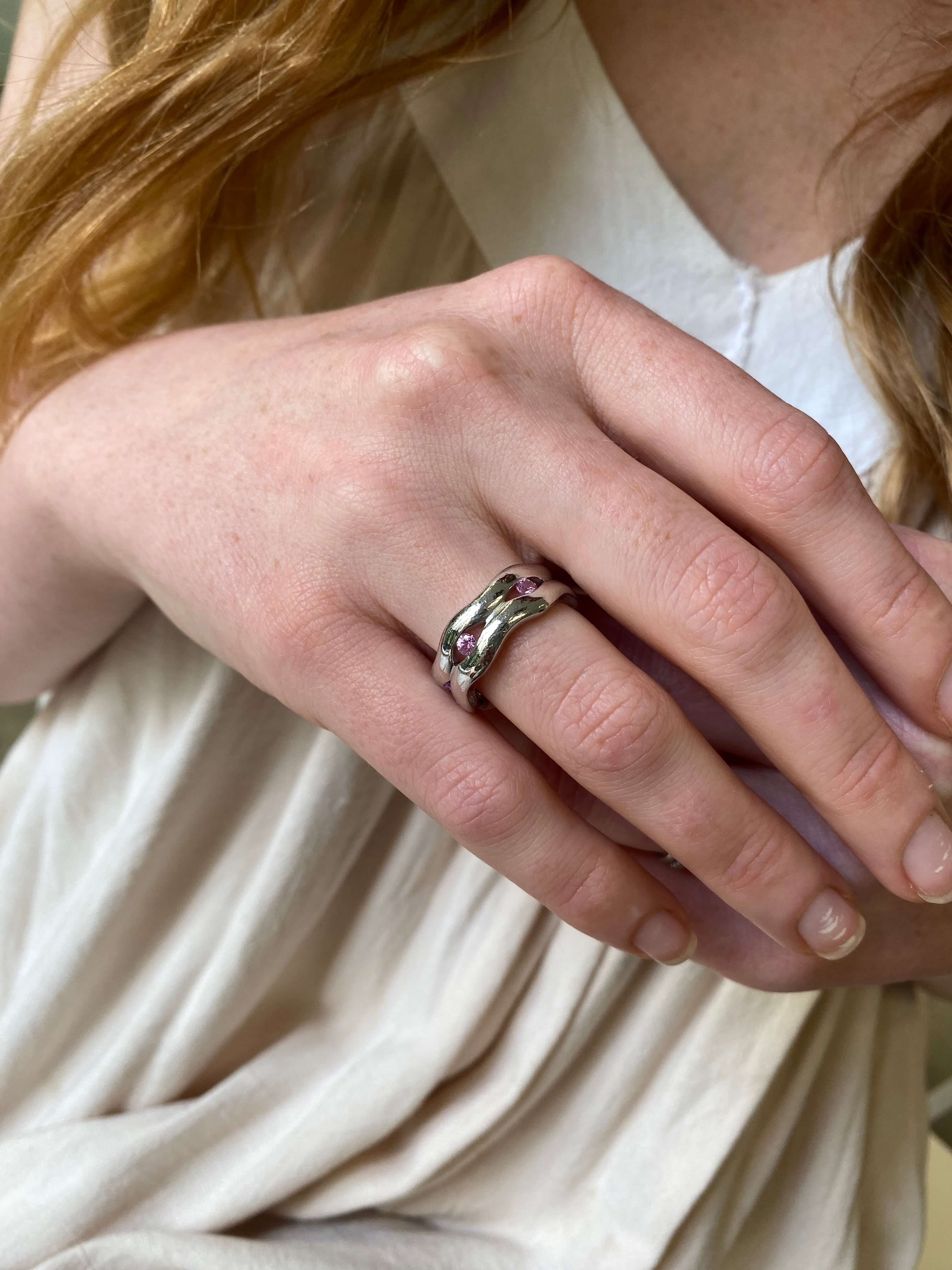 A close-up of a person's hand wearing a silver ring with small pink apphries, set against a soft cream-colored top.