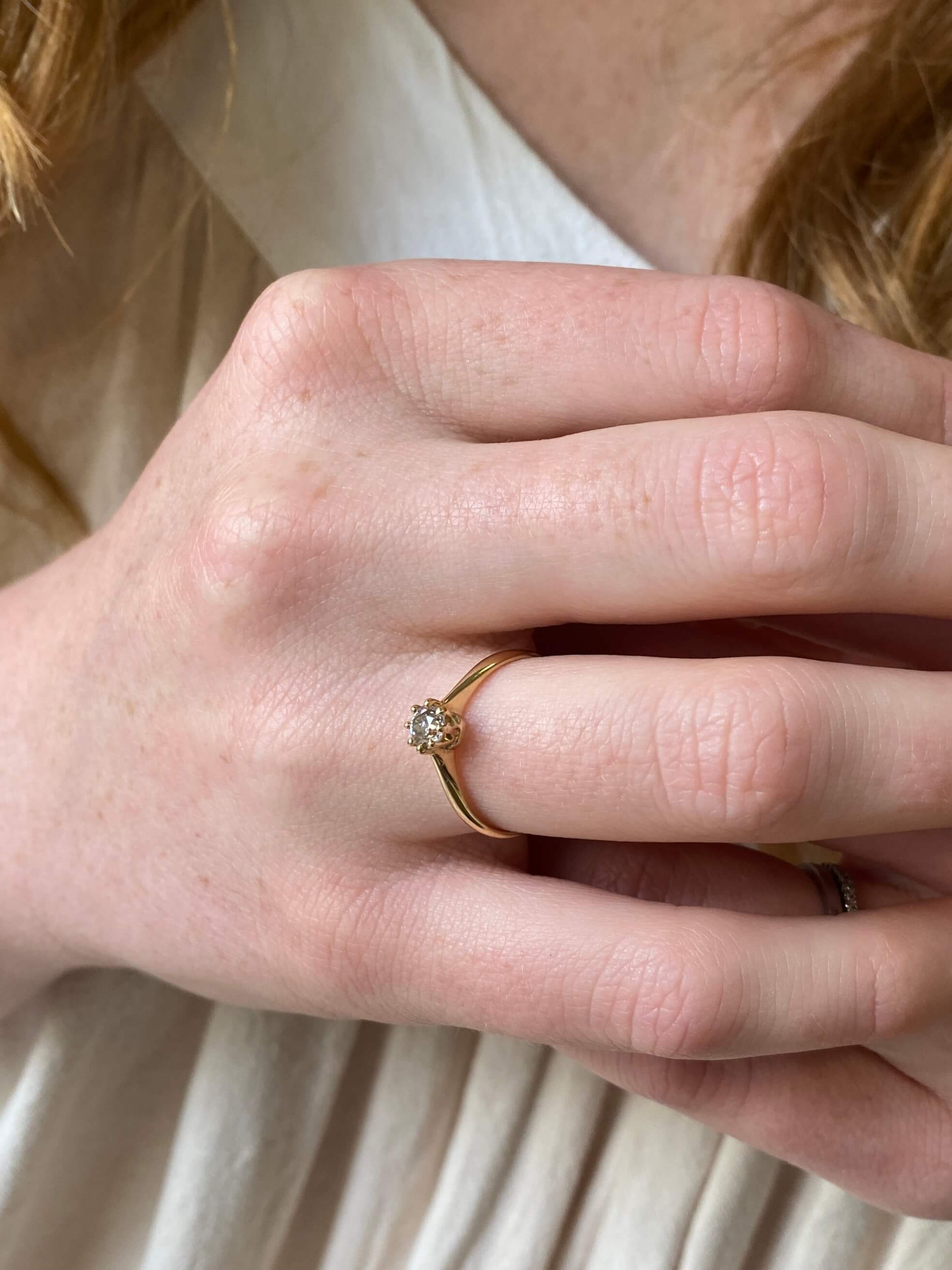 A close-up of a person's hand wearing a delicate yellow gold ring with a small round clear gemstone, set against a soft cream-colored top.