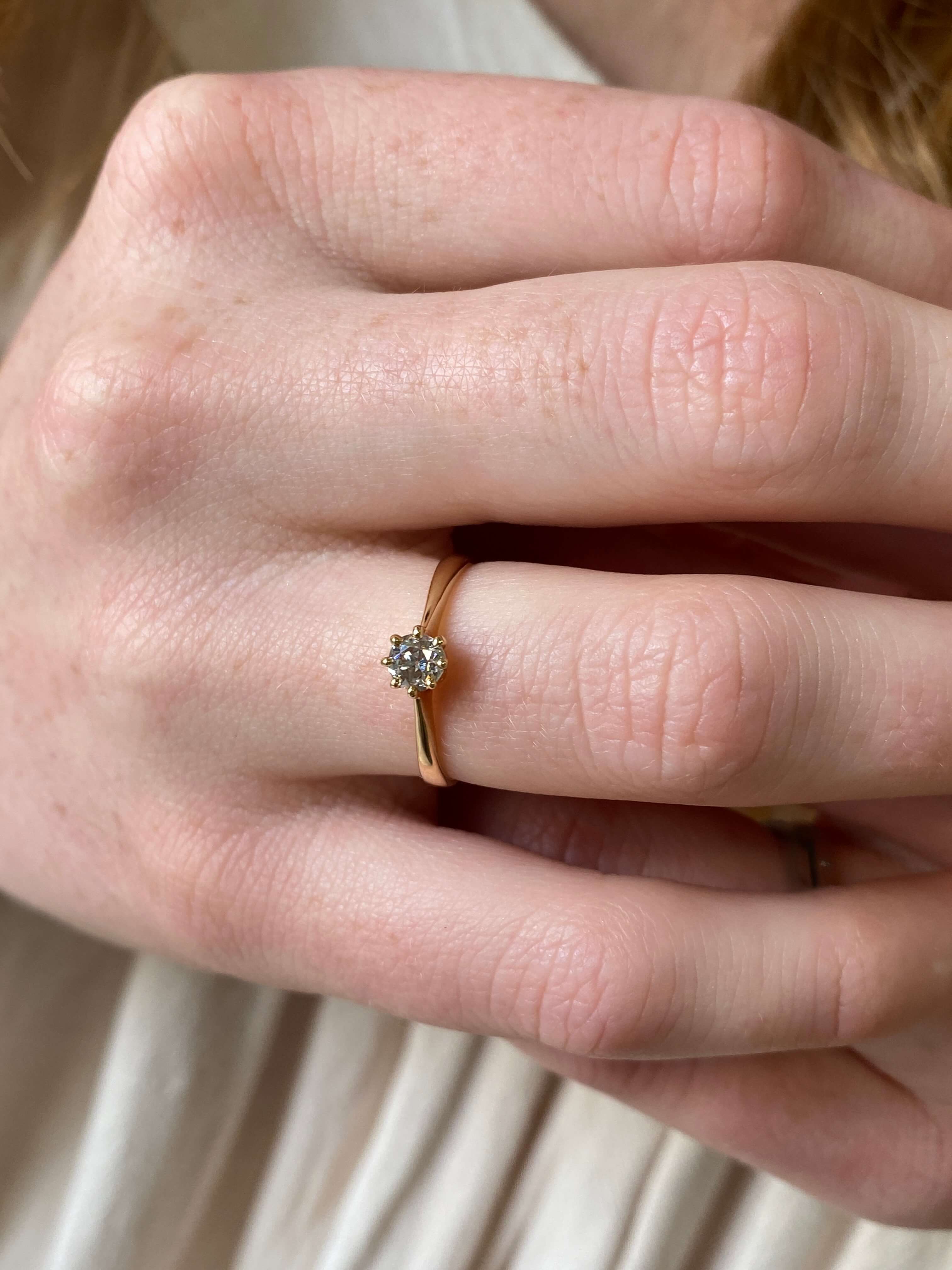 A close-up of a person's hand wearing a delicate yellow gold ring with a single round-cut diamond in a prong setting.