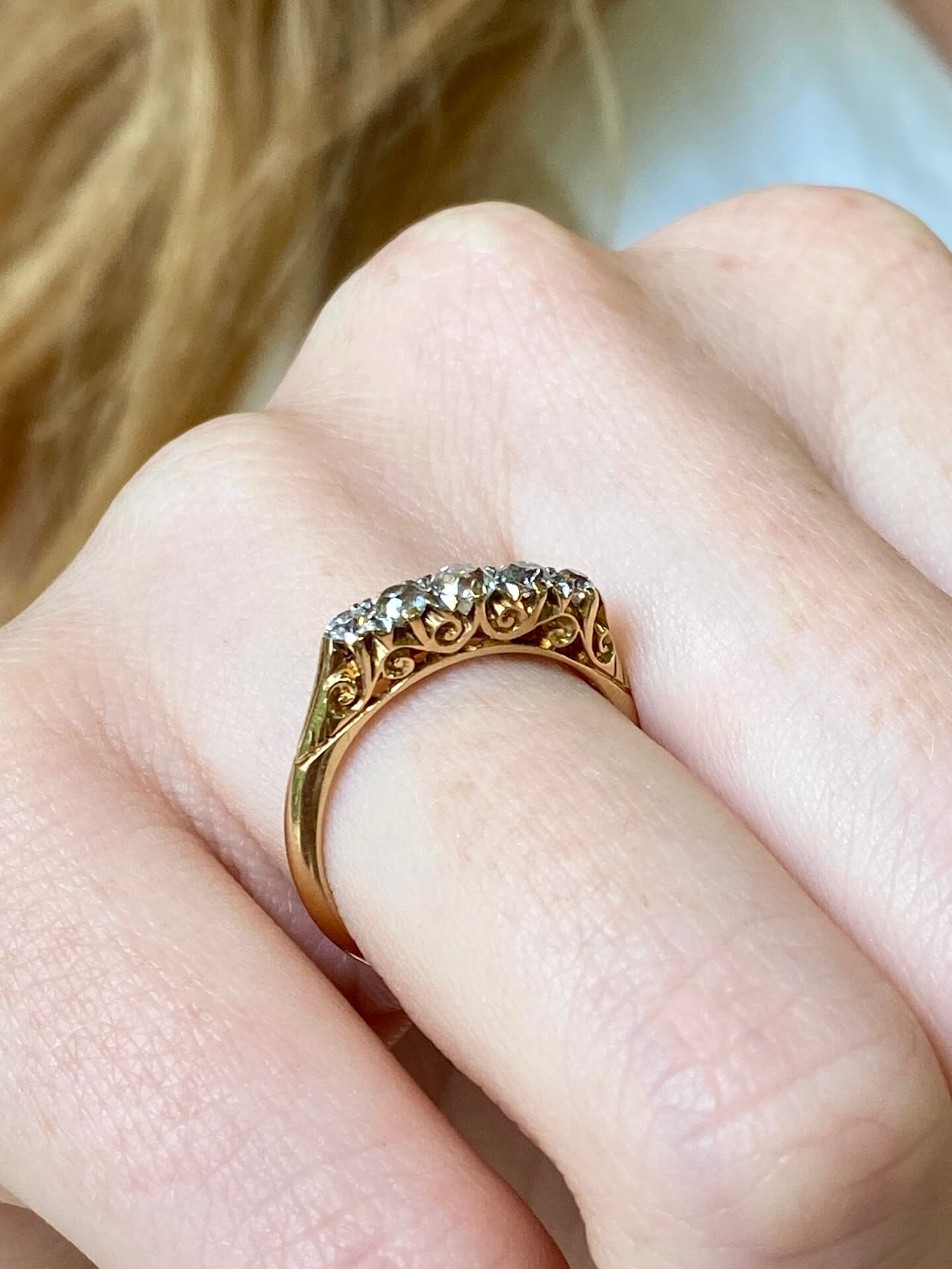 A close-up of a gold ring with a line of old cut diamonds and ornate filigree detailing, worn on a person's finger.