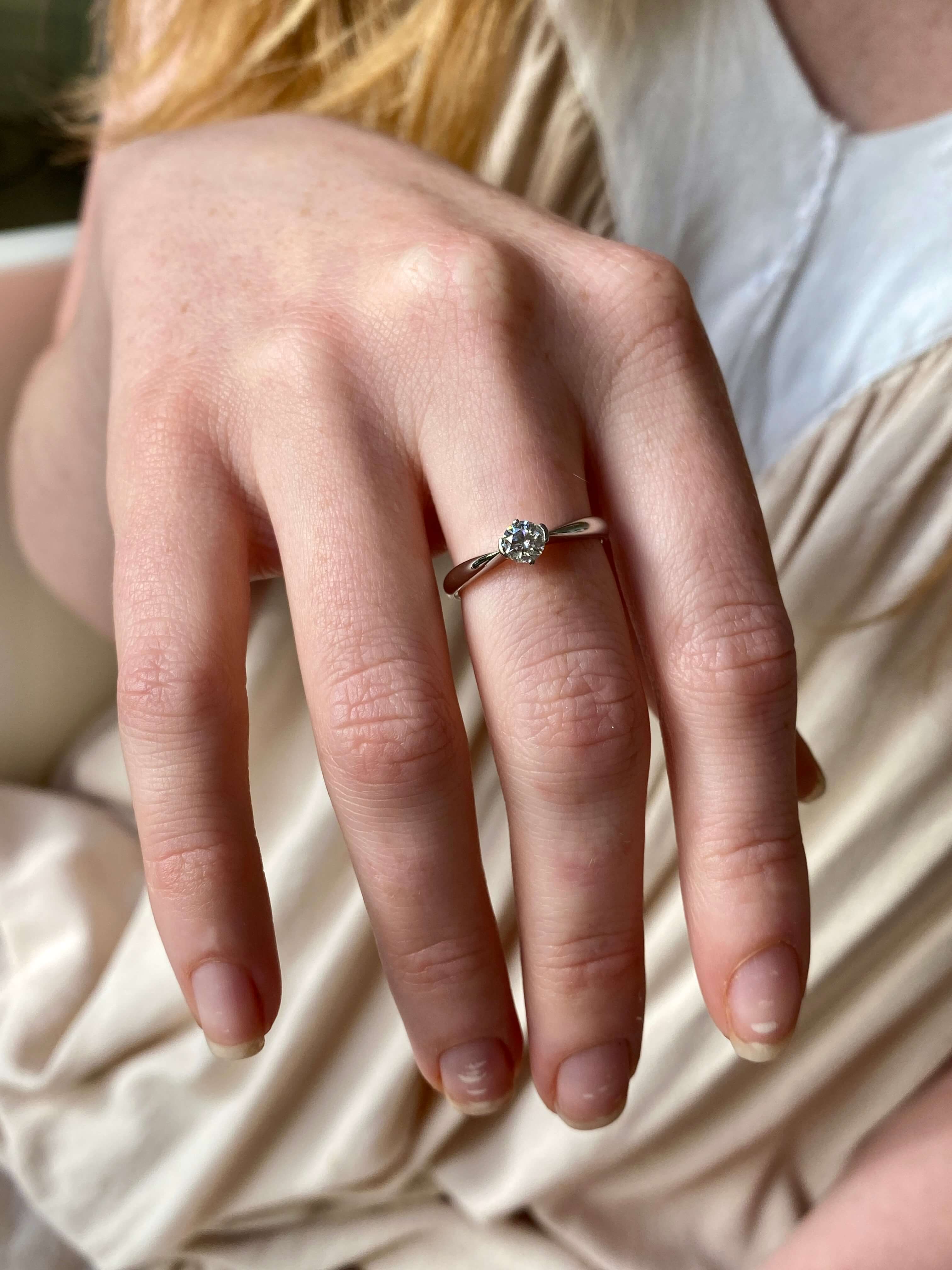 A close-up of a person's hand wearing a delicate silver ring with a single diamond set against a soft beige and white garment background.