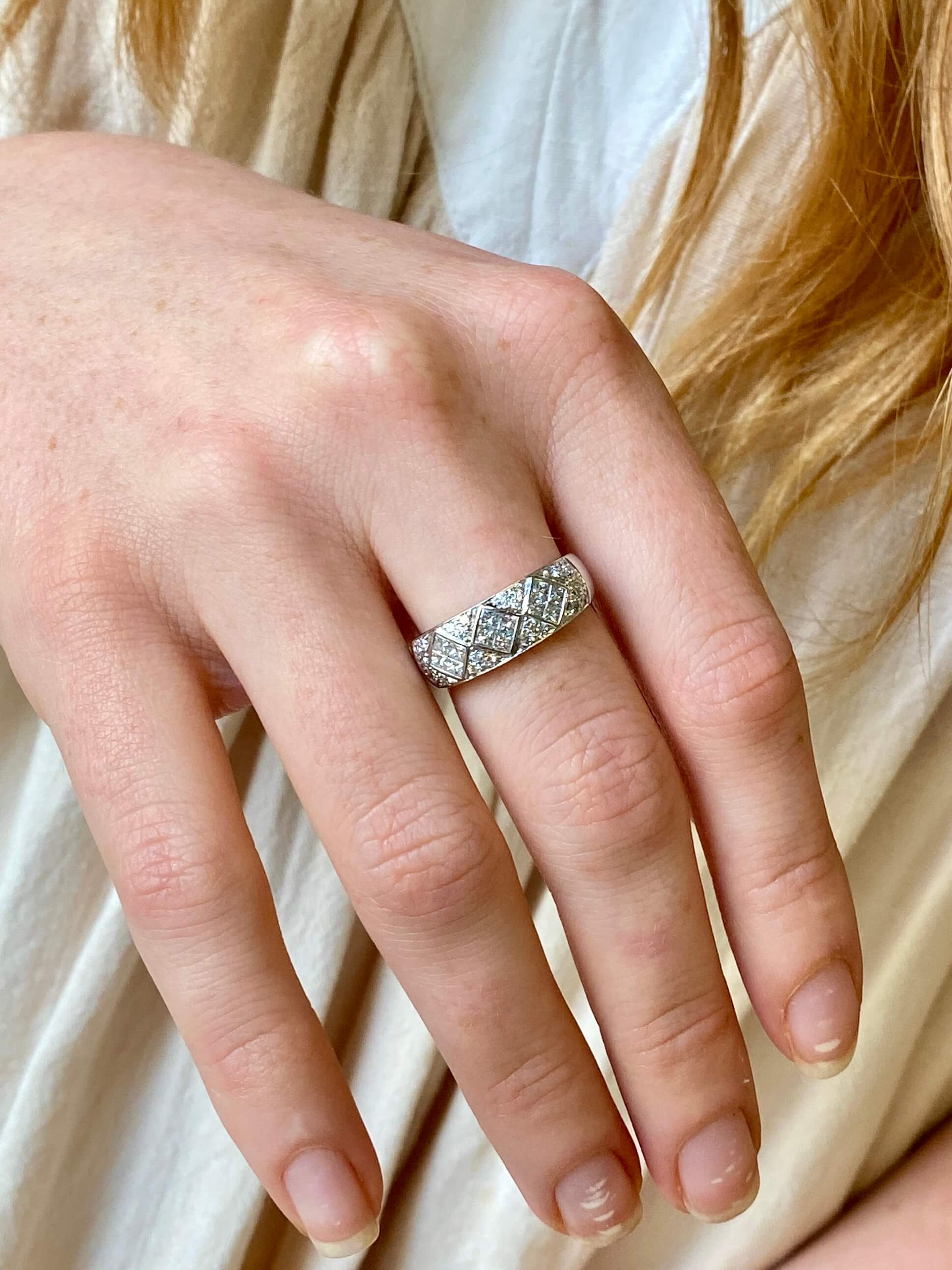 A wide white gold band ring set with square and round diamonds and worn on a hand against a cream dress