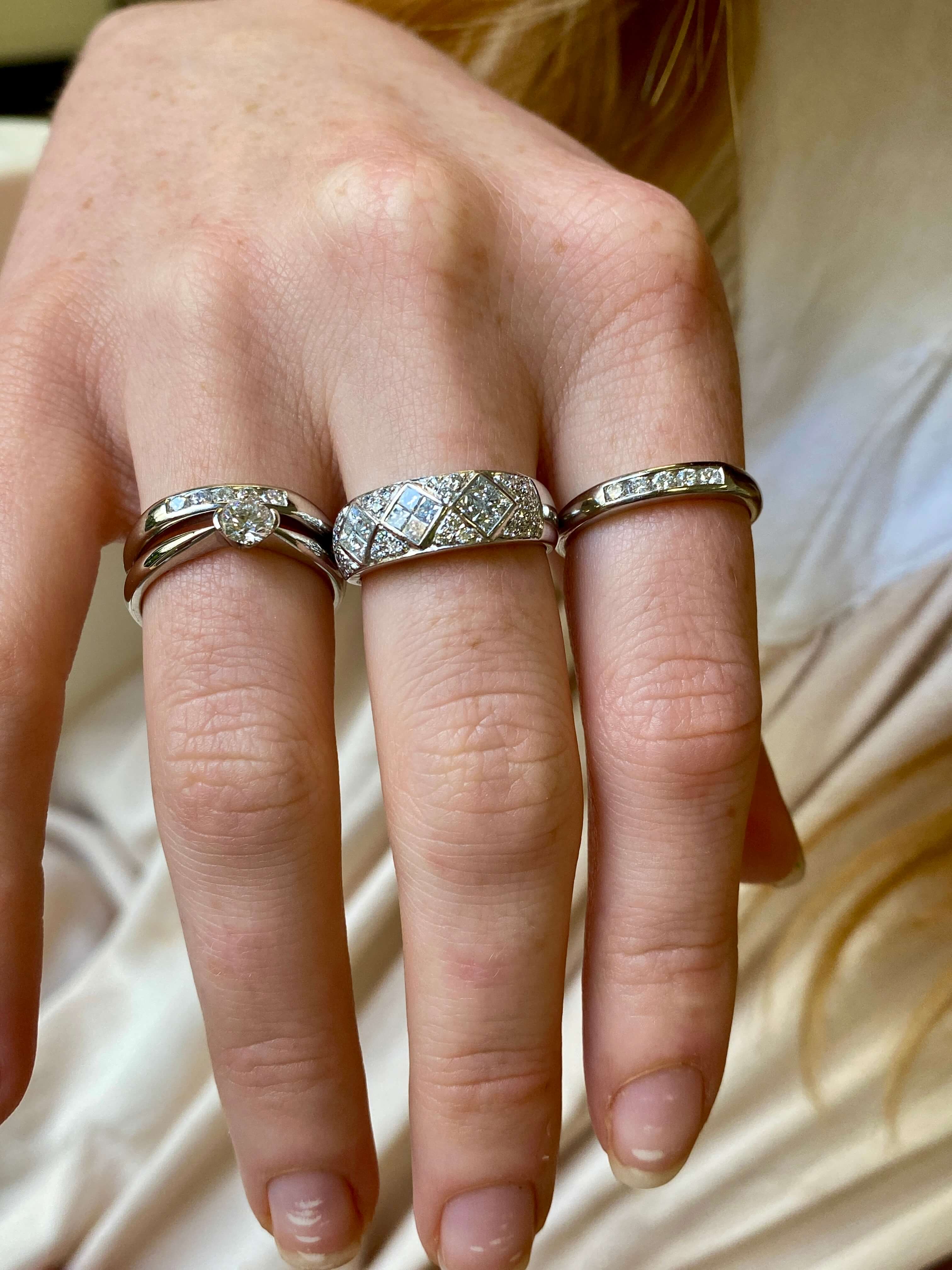 A close-up of a hand wearing multiple rings: a silver band with a round-cut diamond engagement ring, a silver geometric diamond-pattern band, and a silver diamond-accented wedding band, set against a soft cream satin background.