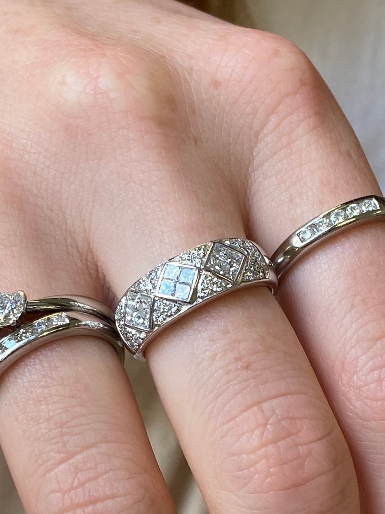 A close-up of a hand wearing a silver-toned ring with a geometric diamond pattern and small clear stones, alongside other rings on the same hand.