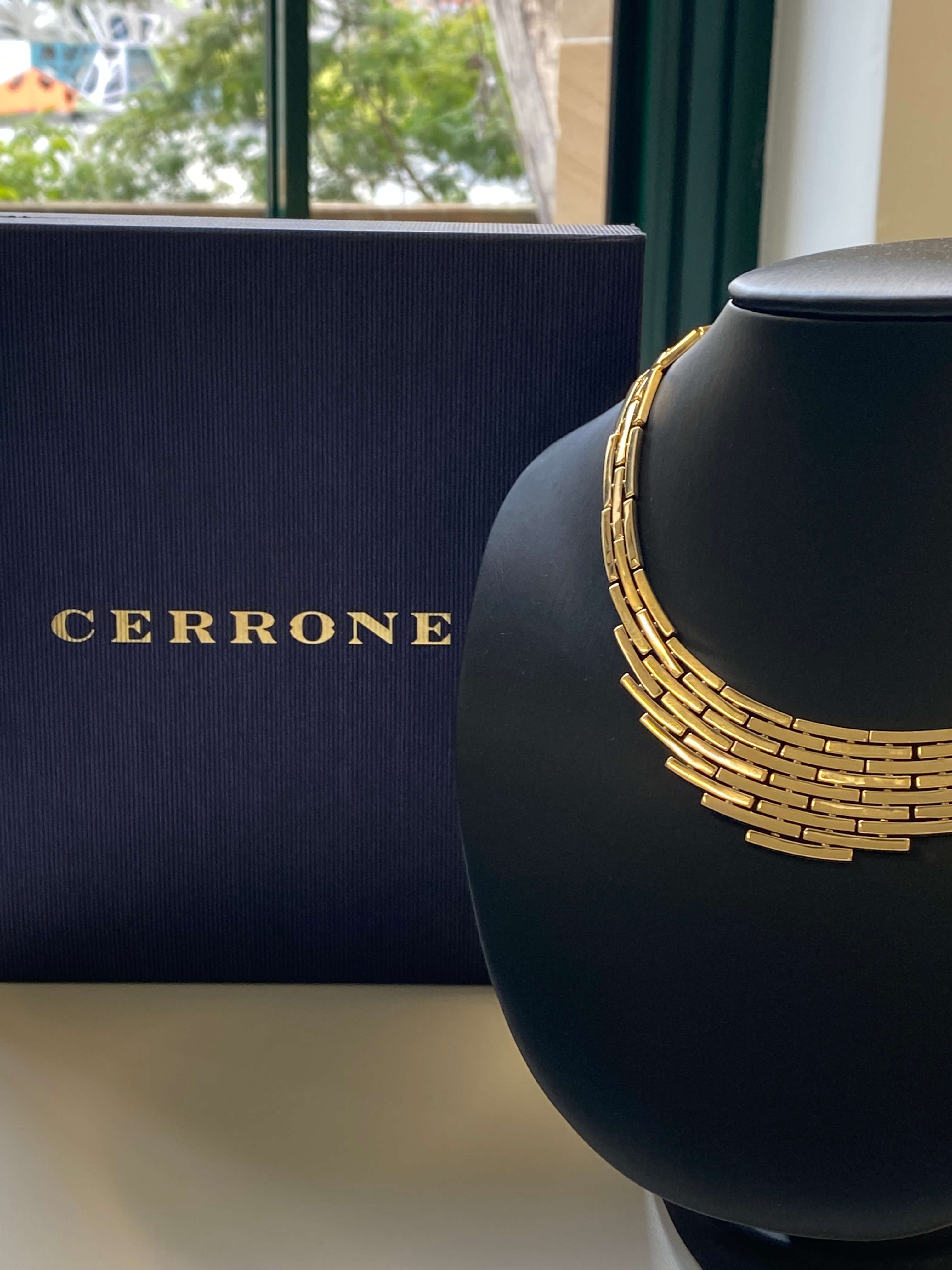A 18ct yellow gold multi-strand collar necklace displayed on a black jewelry bust next to a dark navy blue Cerrone box with gold lettering, set against a softly blurred window background.