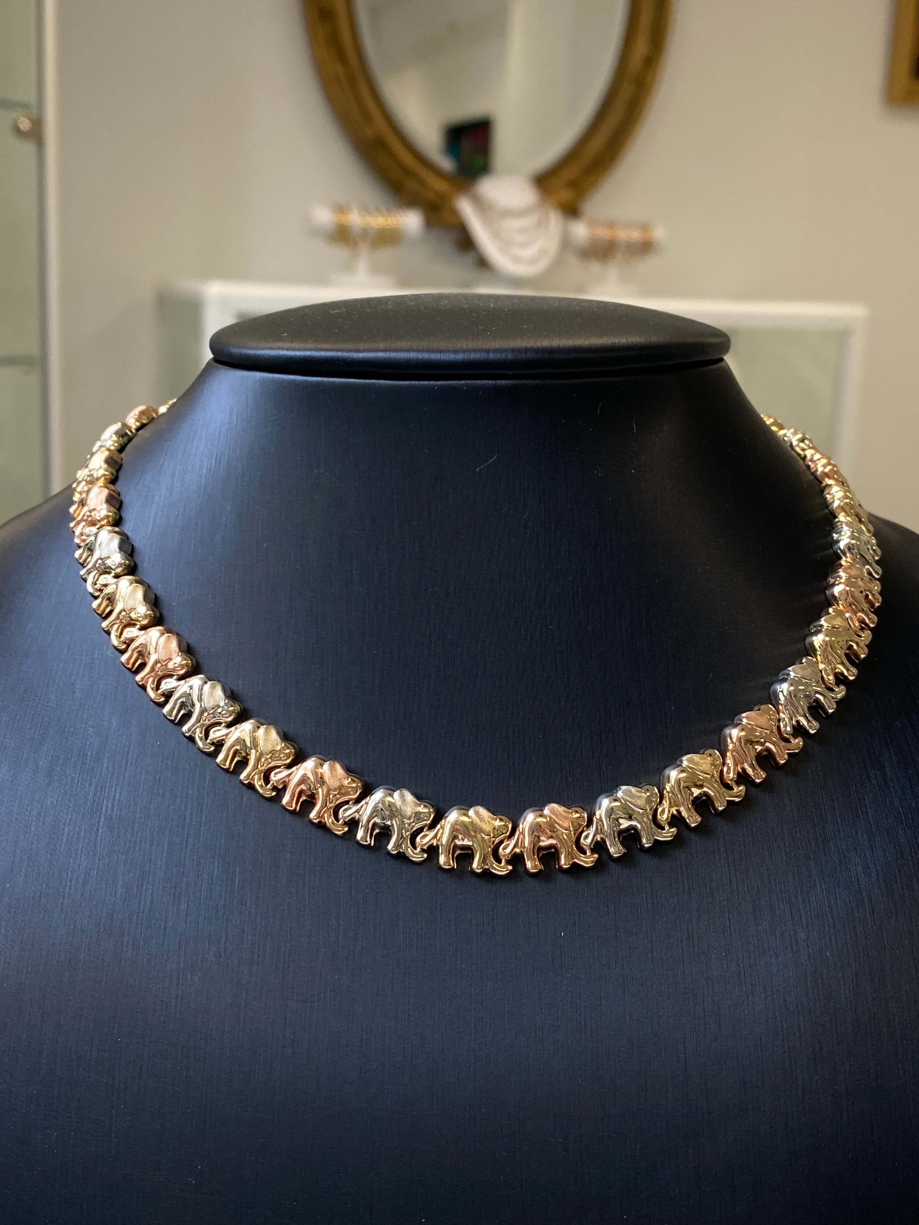 A gold-toned necklace featuring a continuous line of small elephant charms, displayed on a black mannequin bust in a jewelry store setting.