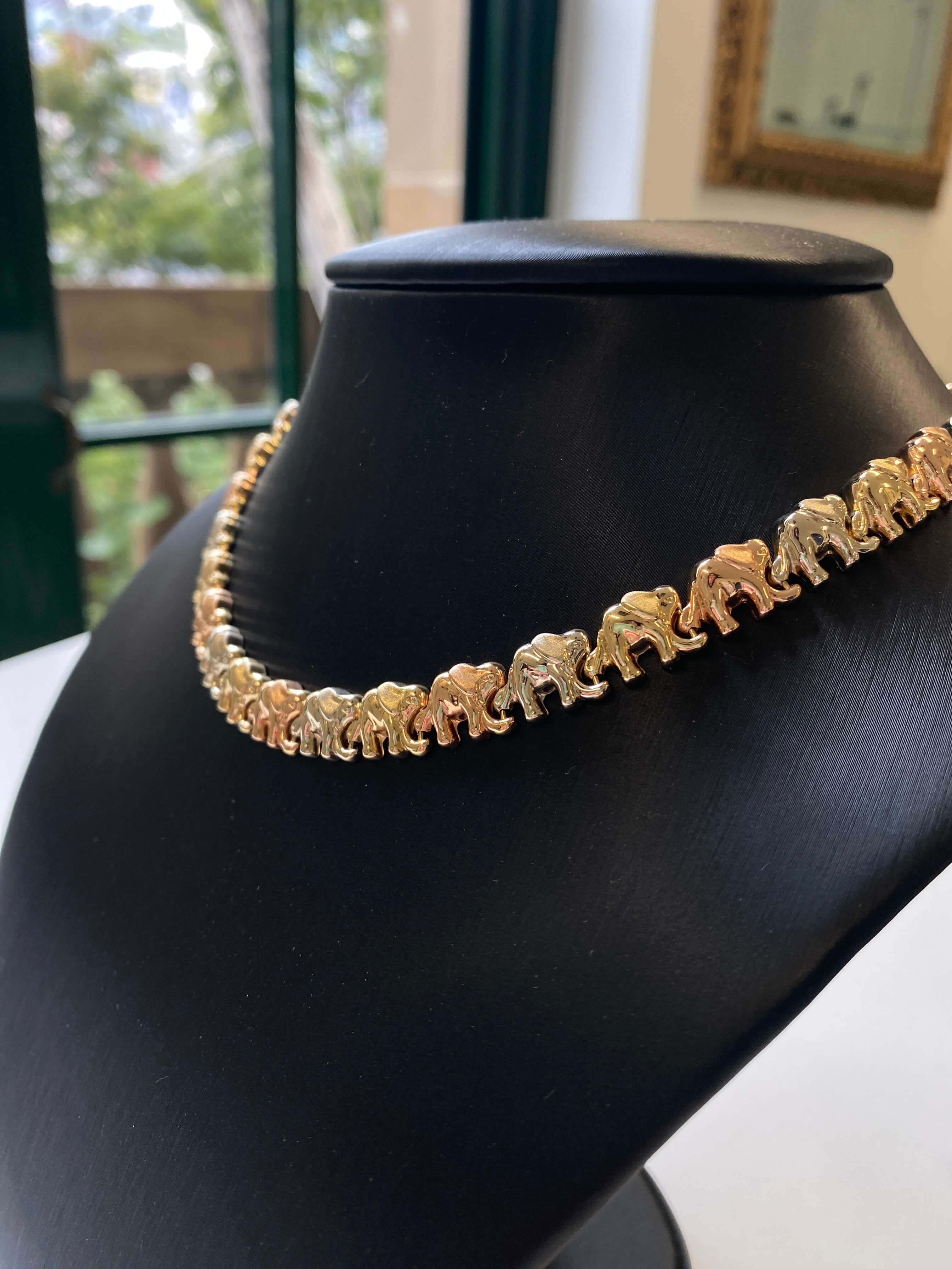 A gold-toned elephant-link chain necklace displayed on a black jewelry bust, with a softly blurred indoor background visible behind it.