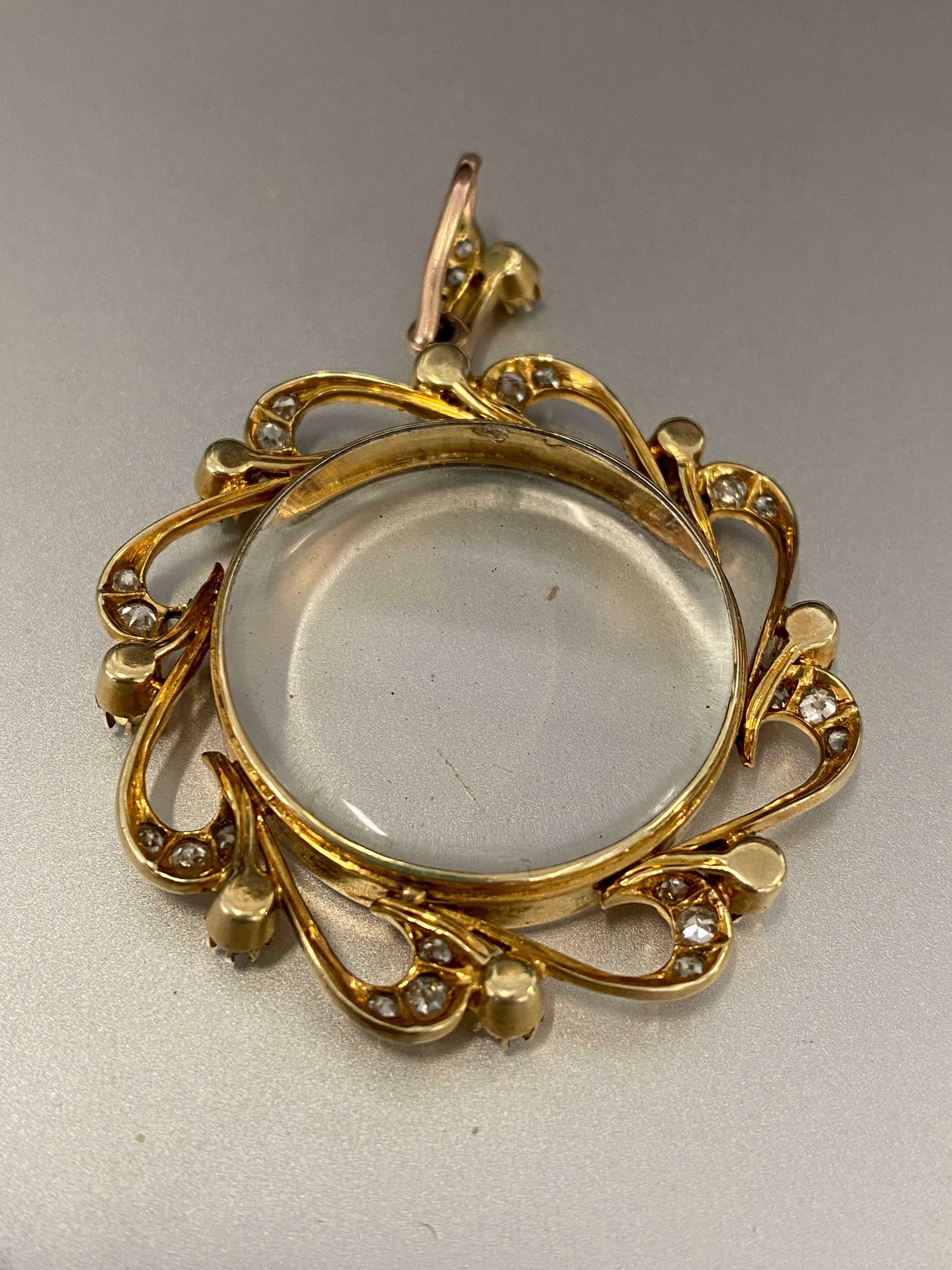 An antique gold photo locket pendant with an ornate scrollwork border and a clear oval center, photographed on a plain light gray background.