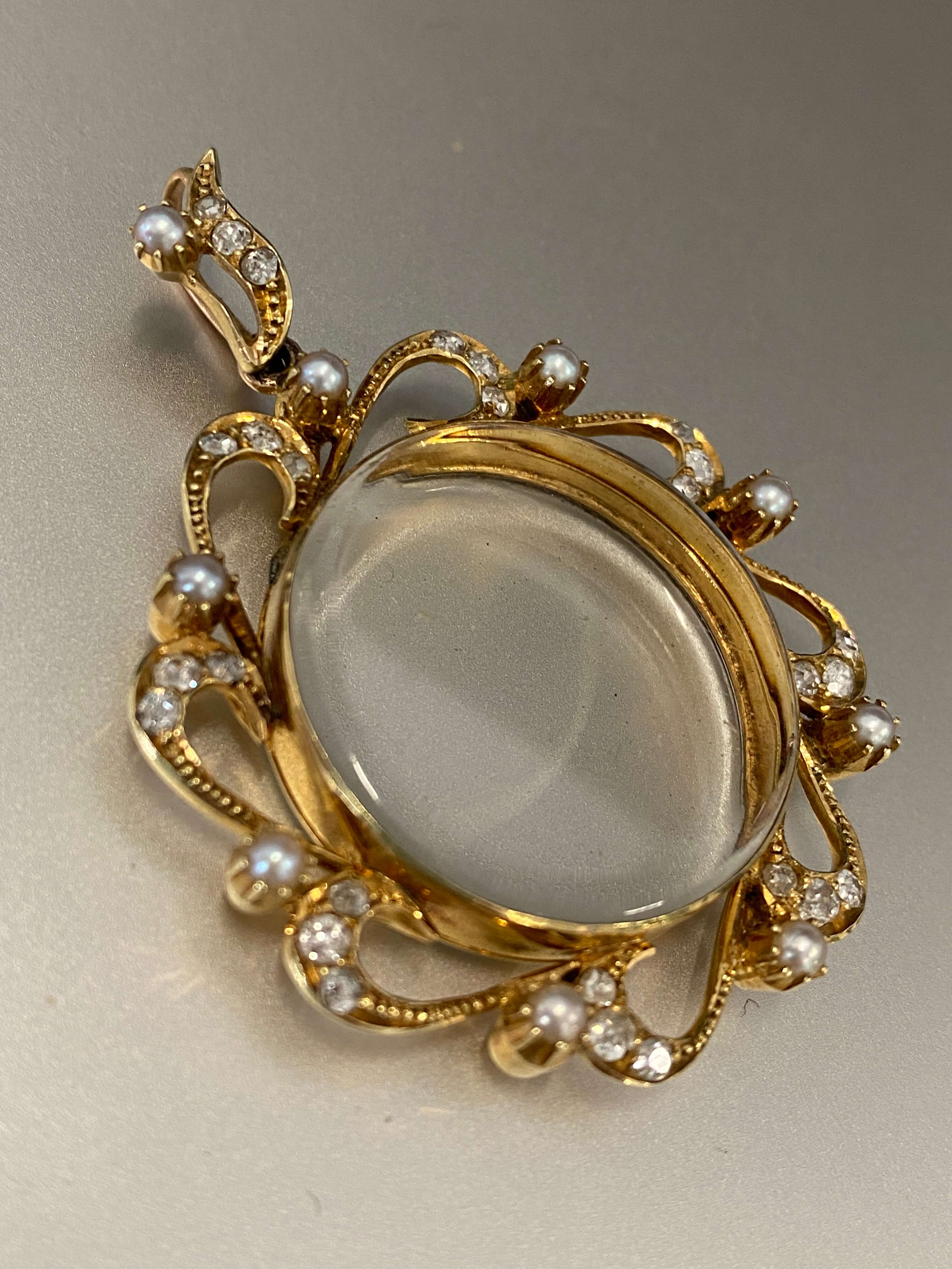 An antique gold oval photo locket featuring clear glass surrounded by ornate gold scrollwork decorated with pearls and diamonds shown on a neutral gray background.