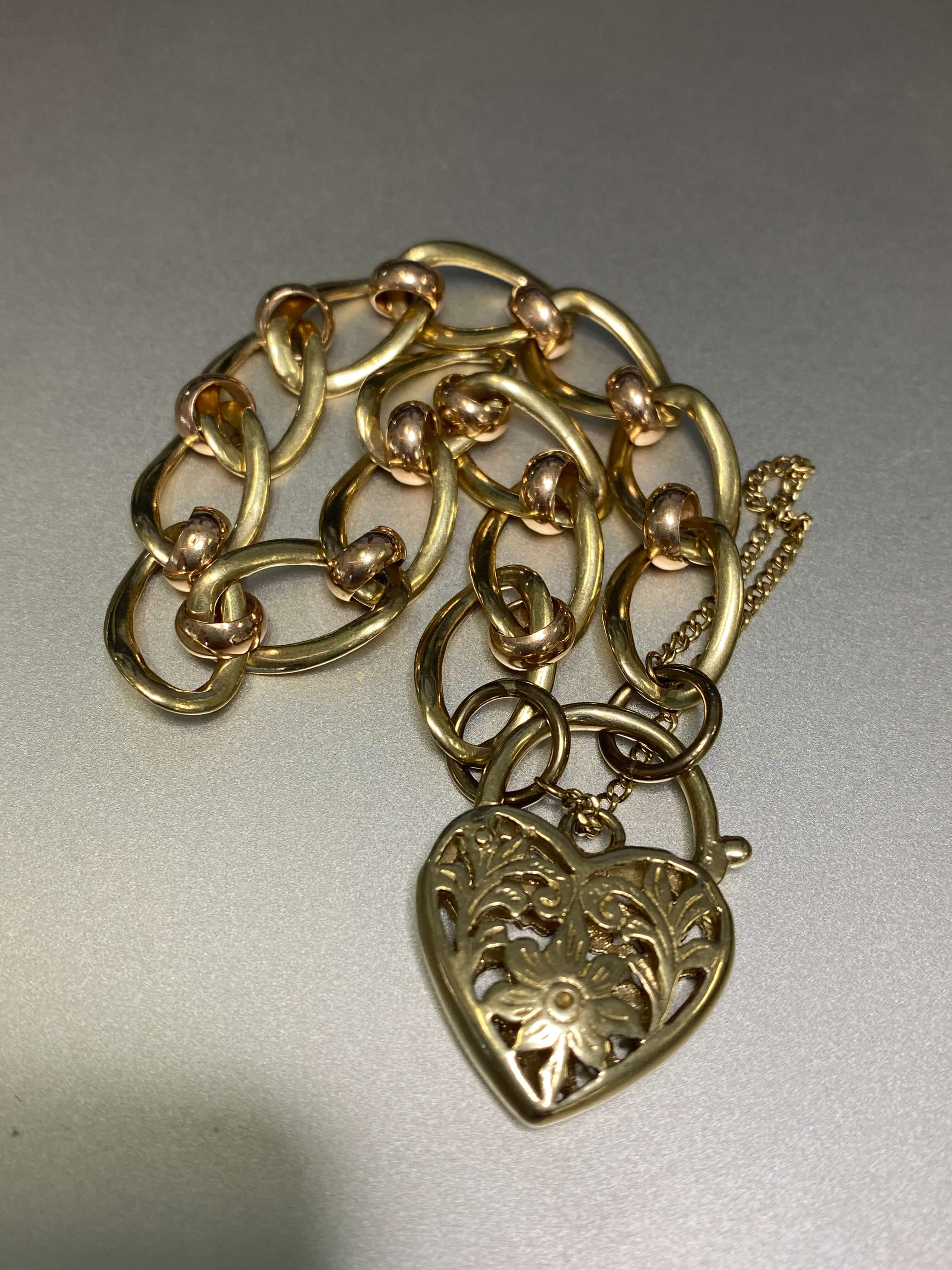A yellow and rose gold bracelet with a heart-shaped filigree padlock clasp, photographed on a plain light gray surface.
