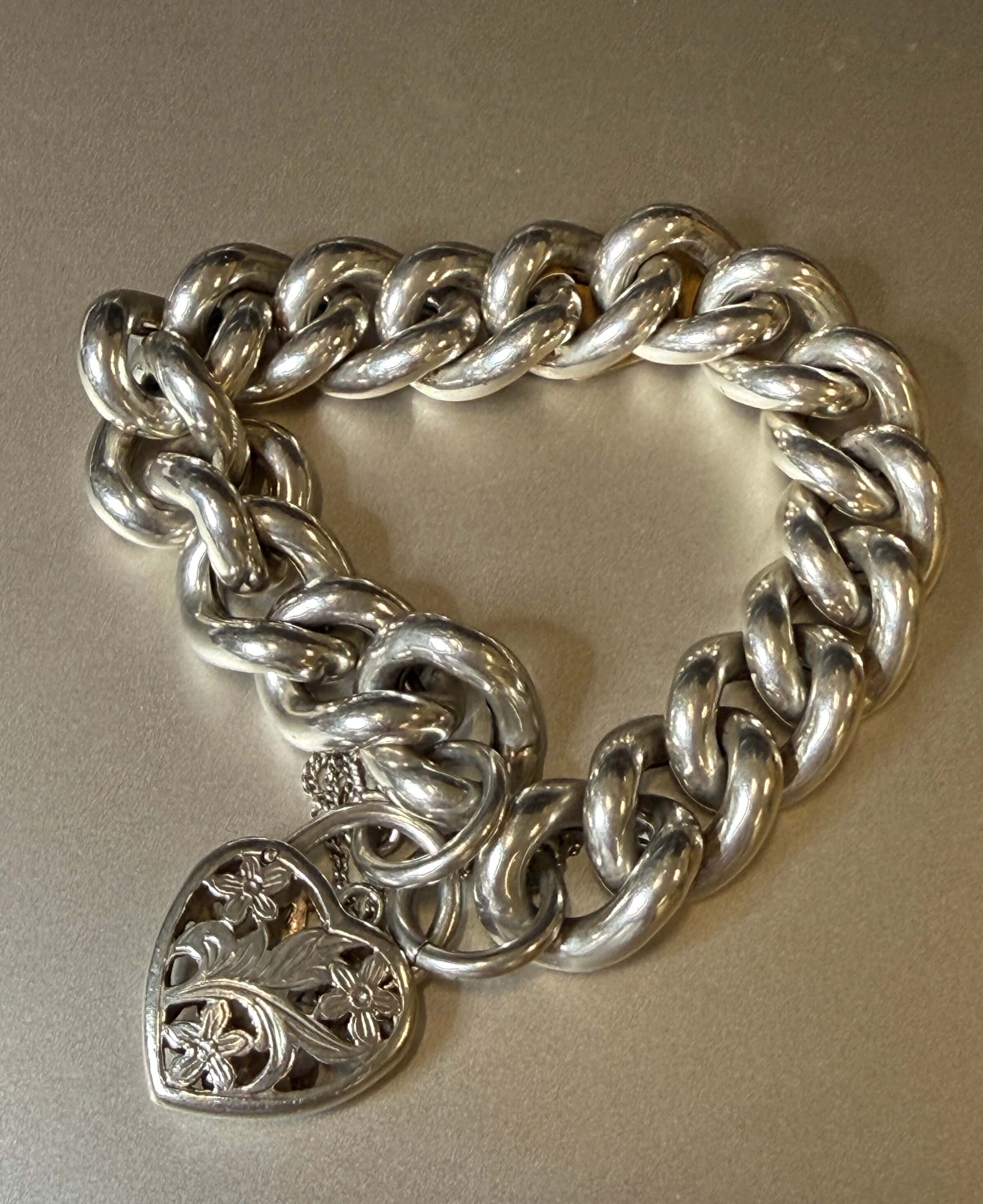 A chunky silver chain bracelet with a heart-shaped filigree pendant, displayed on a neutral beige background.