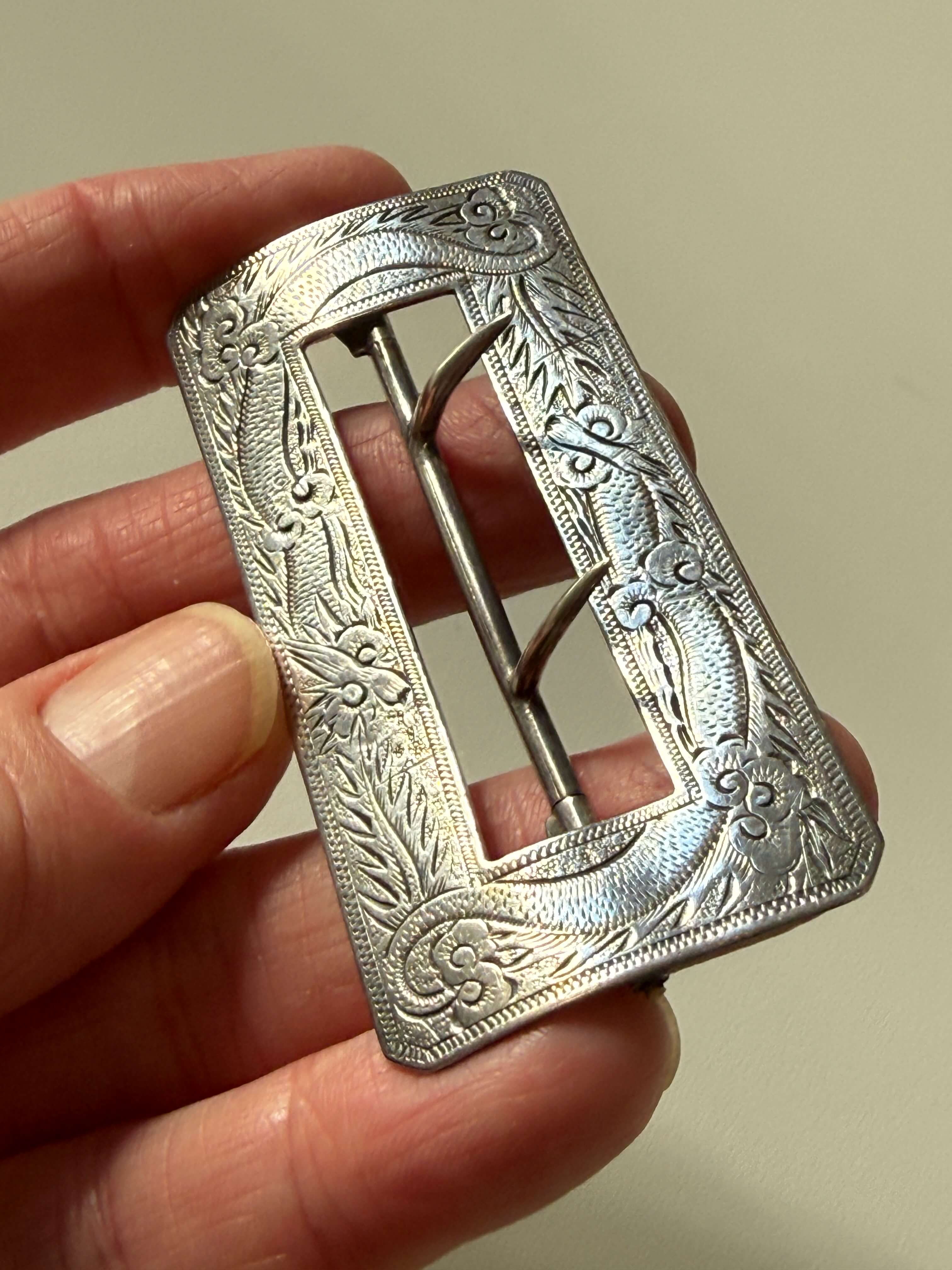 An ornate silver-toned metal buckle with floral engravings, held in a person's hand against a plain beige background.