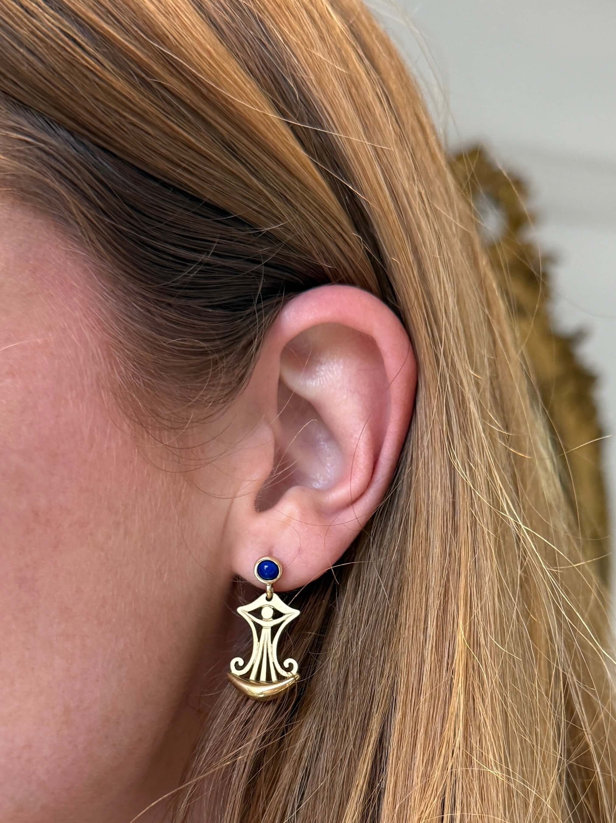 A close-up of a person's ear wearing a gold earring with a small dark blue round stud and a dangling gold filigree charm.