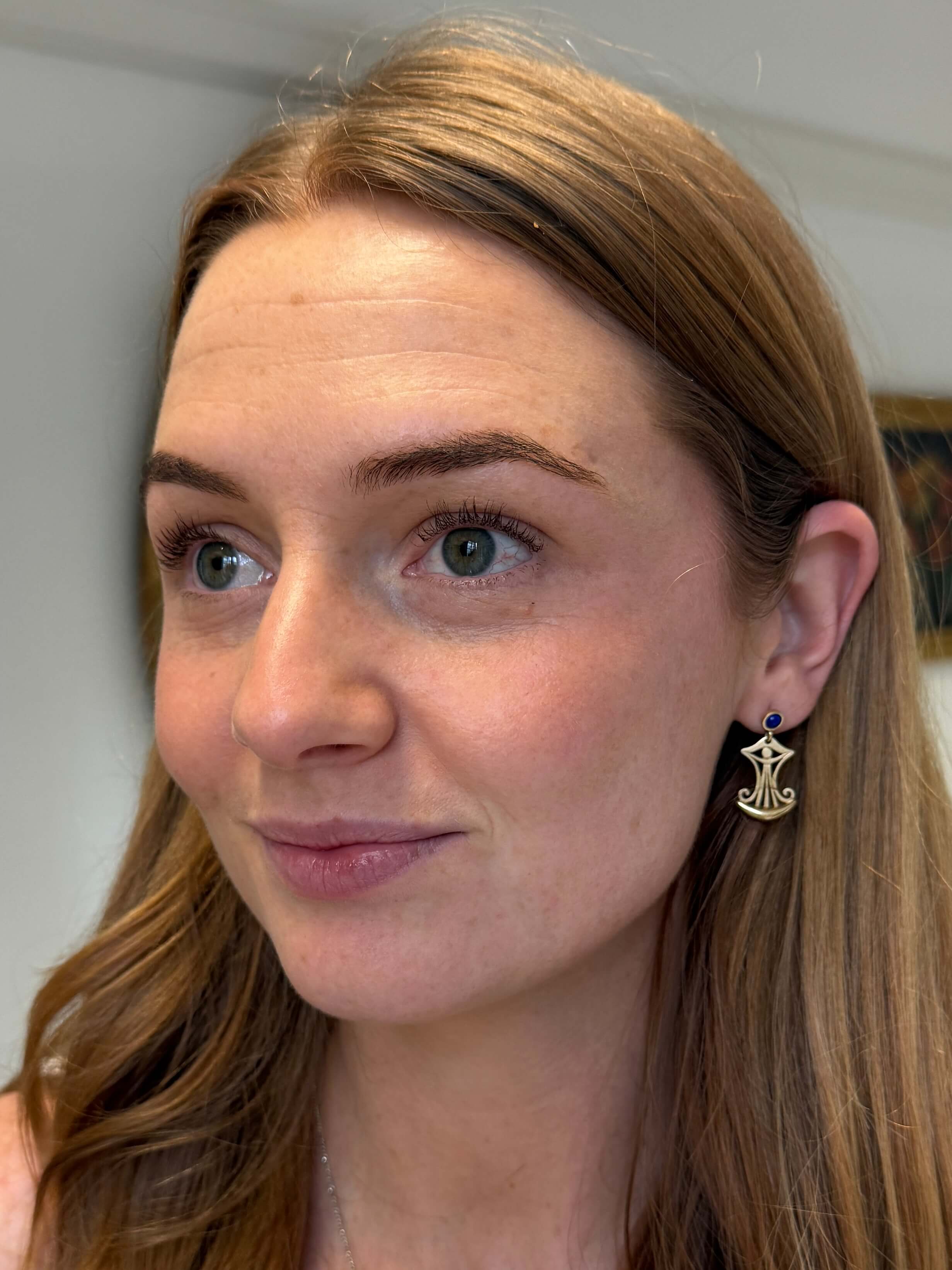 A close-up portrait of a woman with light brown hair wearing a gold anchor-shaped earring with a small blue gemstone accent.