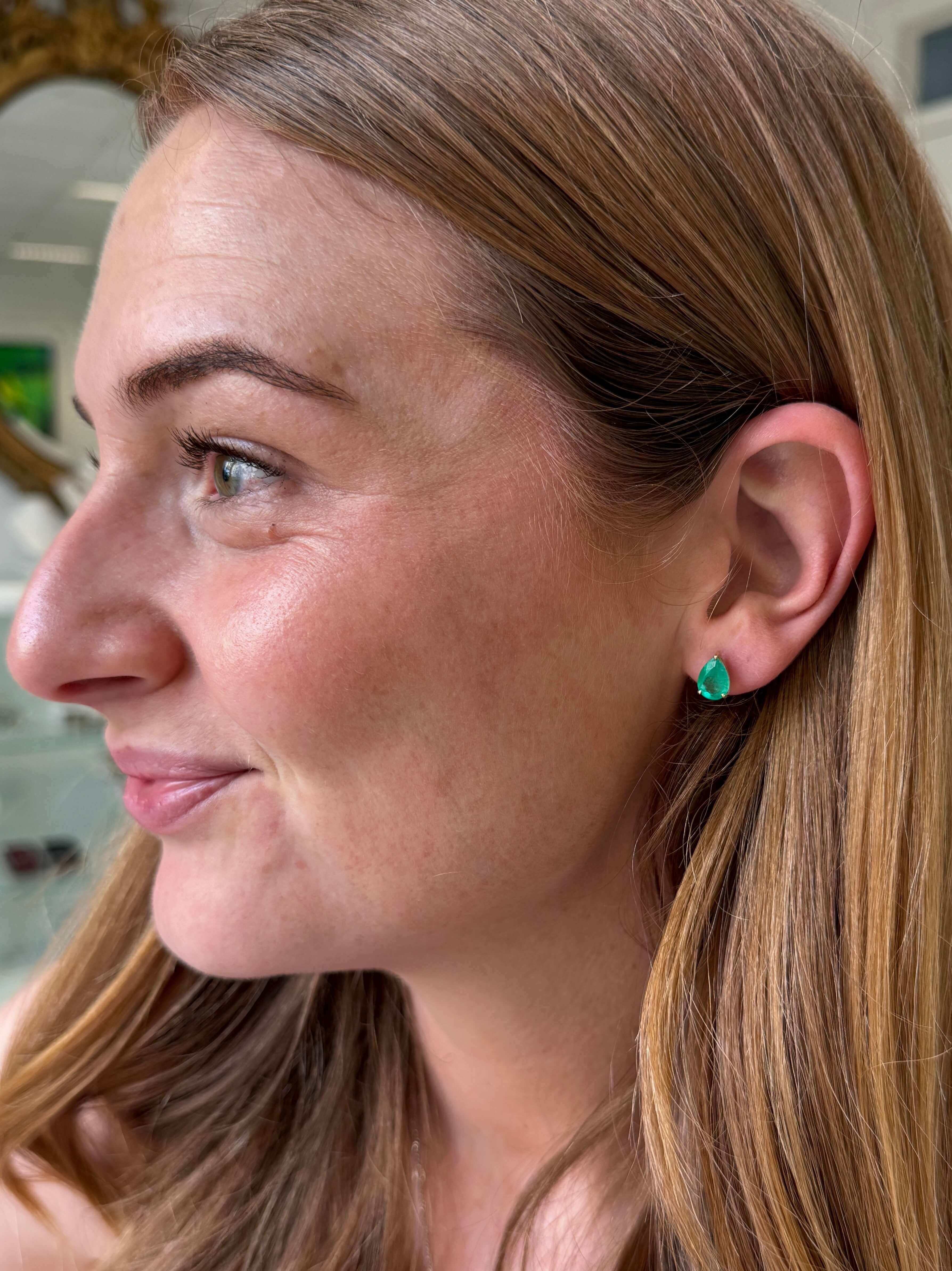 A close-up side profile of a woman wearing wearing a pear-shaped emerald stud earring.