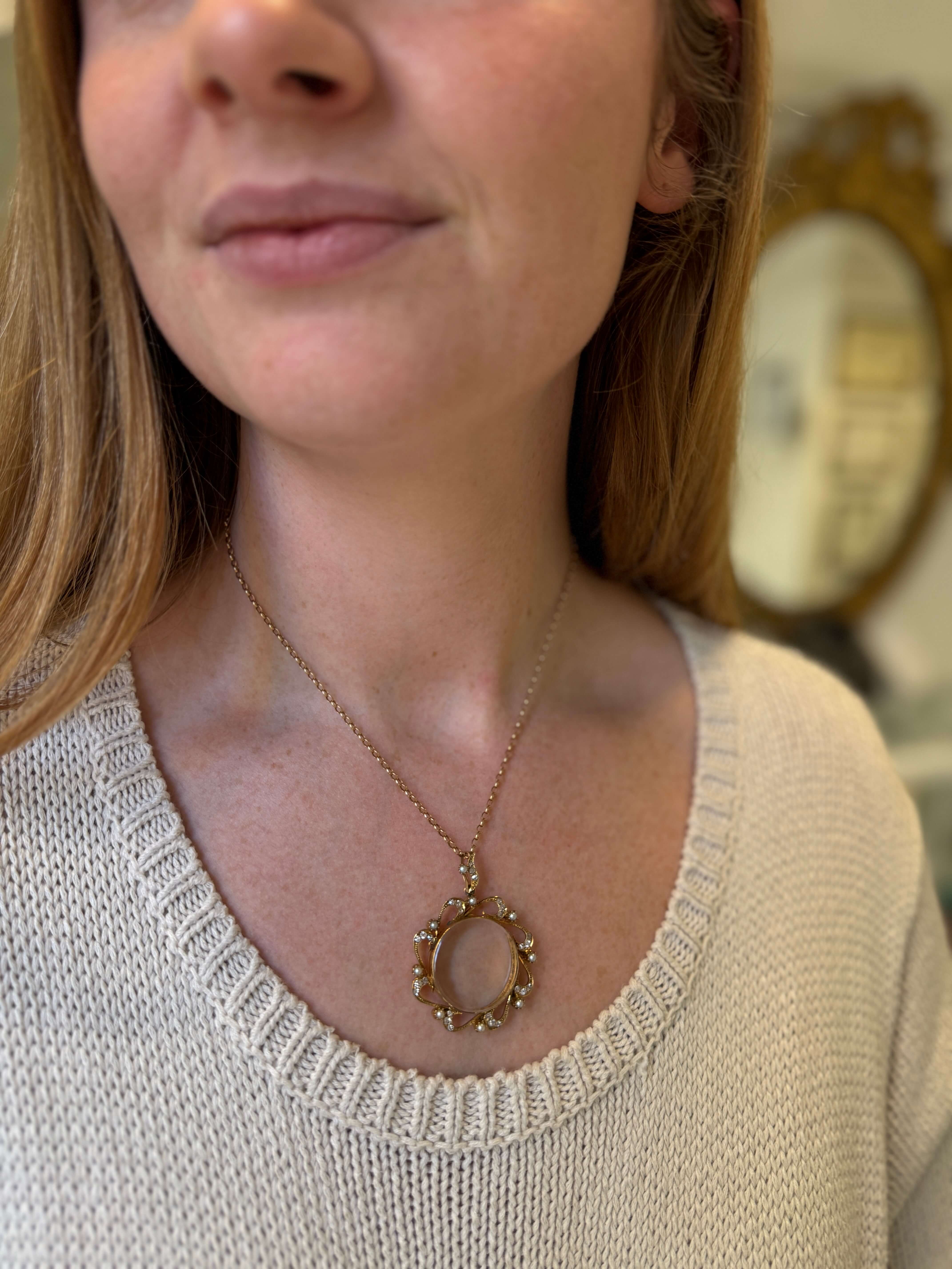 A close-up of a woman wearing a delicate gold necklace with a circular pearl & diamond pendant featuring a pale pink stone in the center, paired with a cream-colored knit sweater.